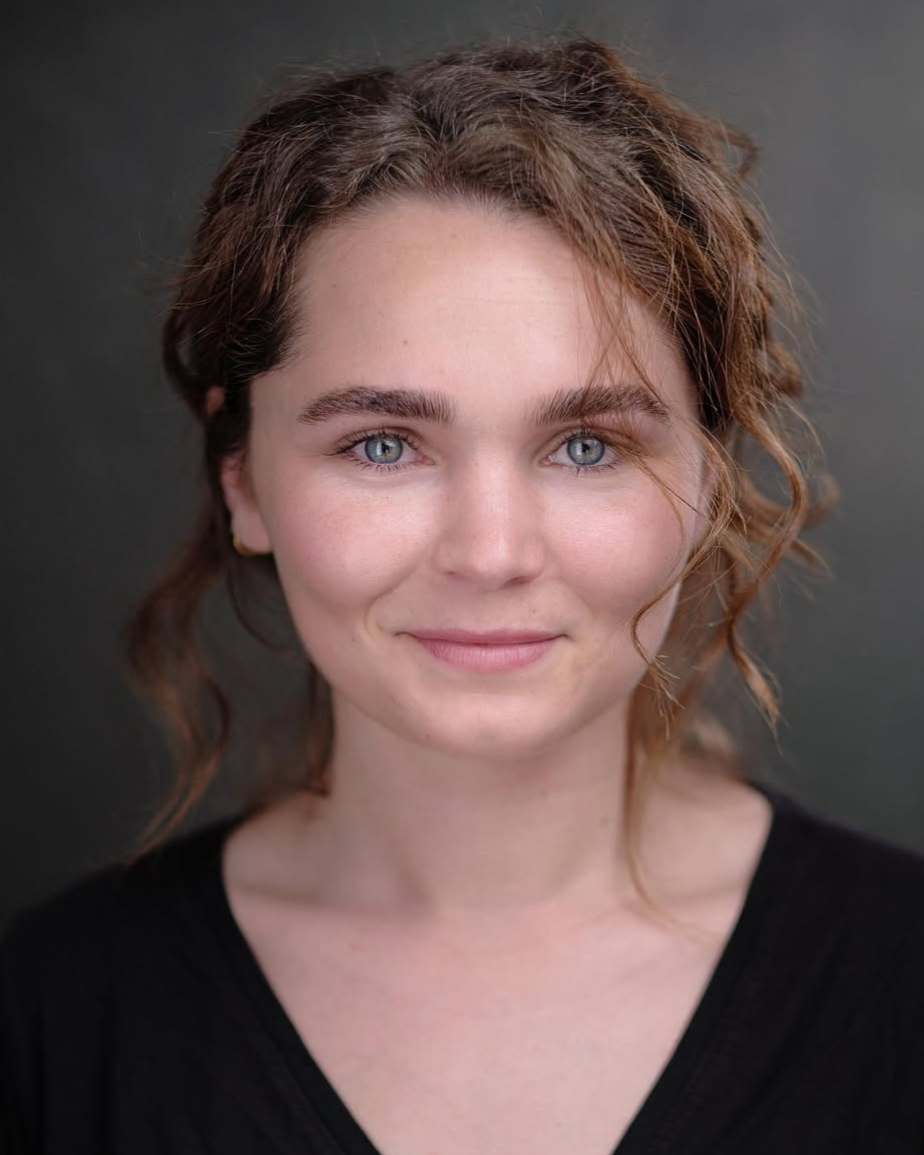 Close-up portrait of a woman with curly brown hair and blue eyes, wearing a black top, against a dark background.