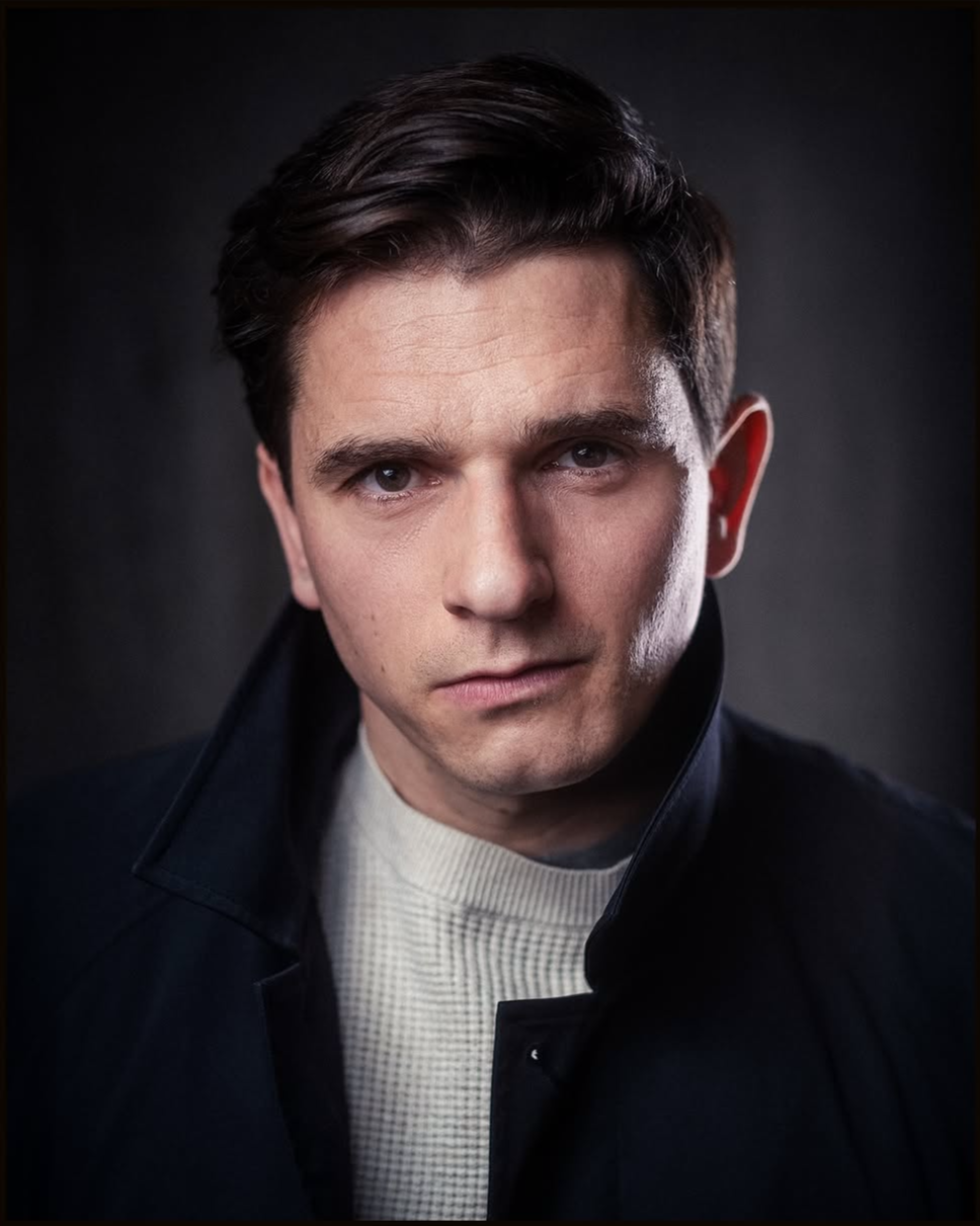 Close-up portrait of a young man with short, dark hair, wearing a black jacket and a light-colored sweater, against a dark background.