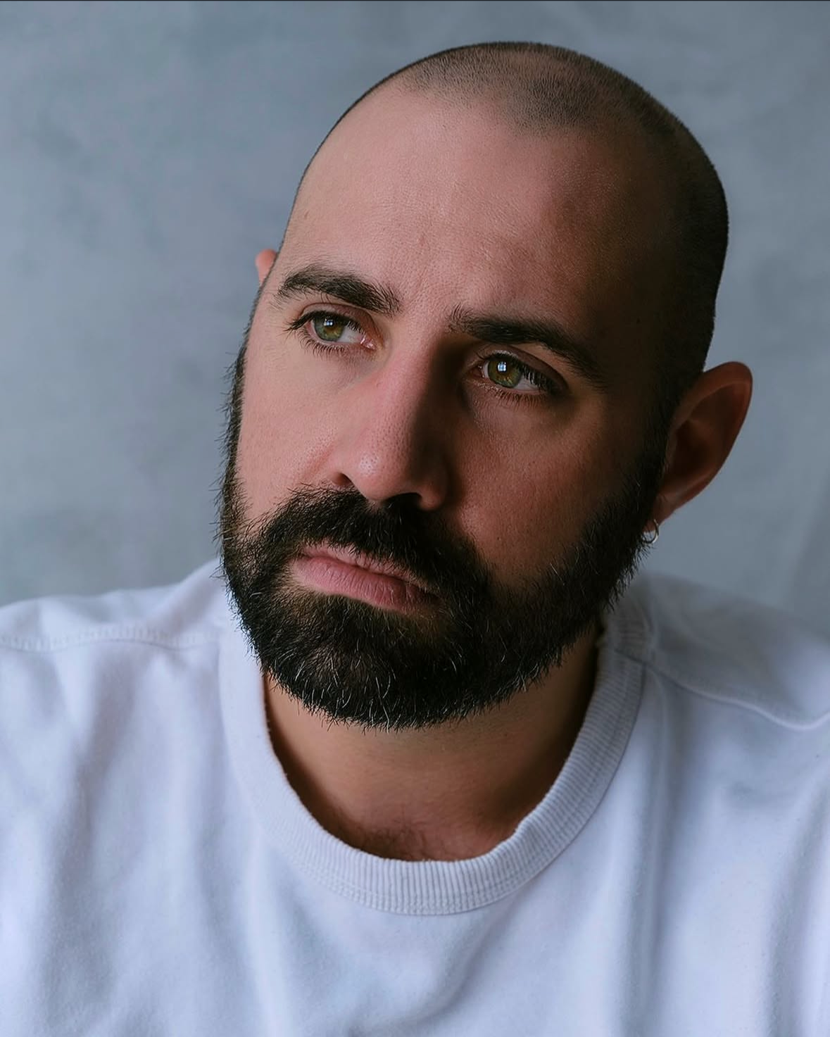 A man with a shaved head and full beard, wearing a white shirt, looking thoughtfully into the distance with a neutral expression.