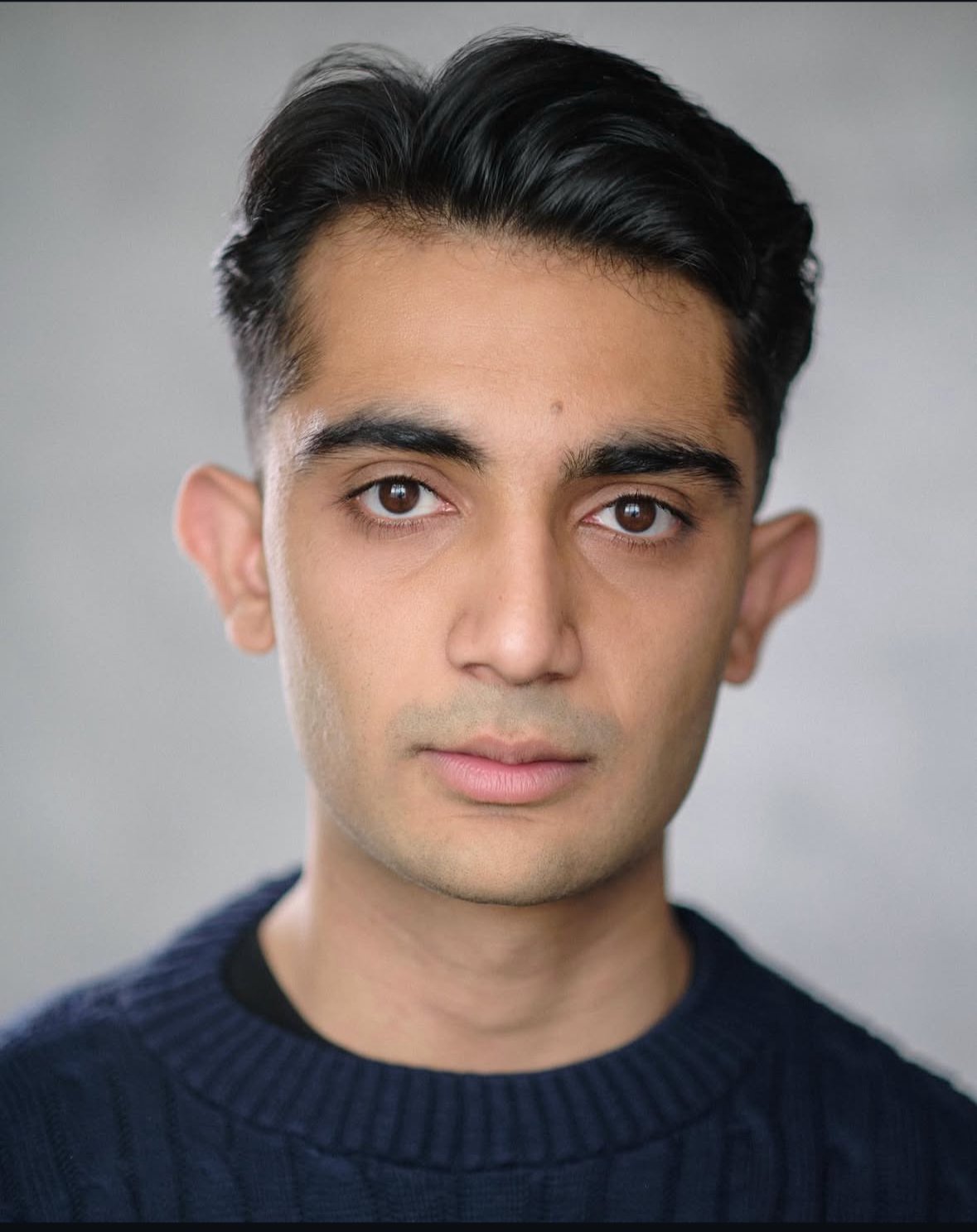 Close-up portrait of a young man with dark hair, brown eyes, wearing a dark blue sweater, against a neutral gray background.