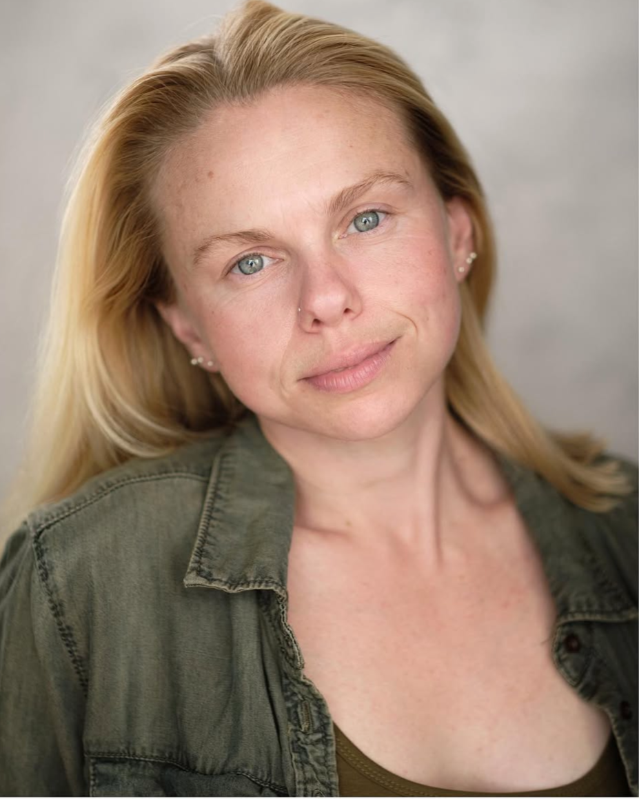 Close-up portrait of a woman with blonde hair, blue eyes, and light skin, wearing a green jacket and a dark top, smiling softly.