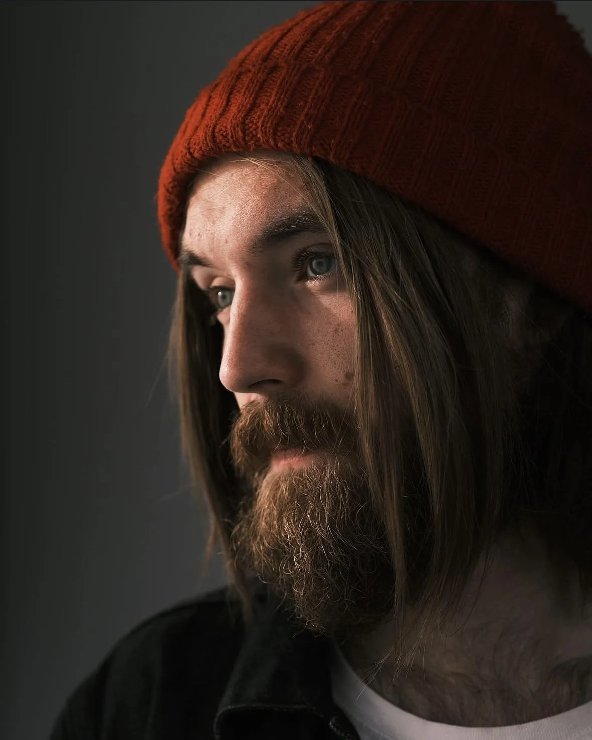 Close-up profile of a man with long hair, a beard, wearing an orange knit beanie, looking to the left.
