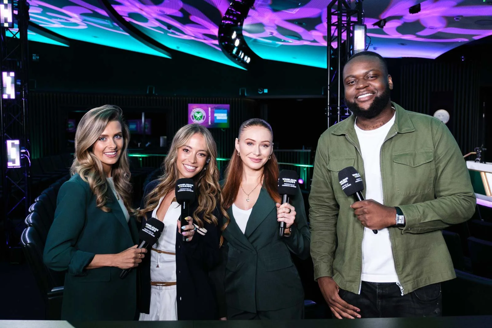 Four people, three women and one man, holding microphones with 'Wimbledon Championships' written on them, standing in a dimly lit room with colorful digital lighting on the ceiling and a green-lit bar area in the background.