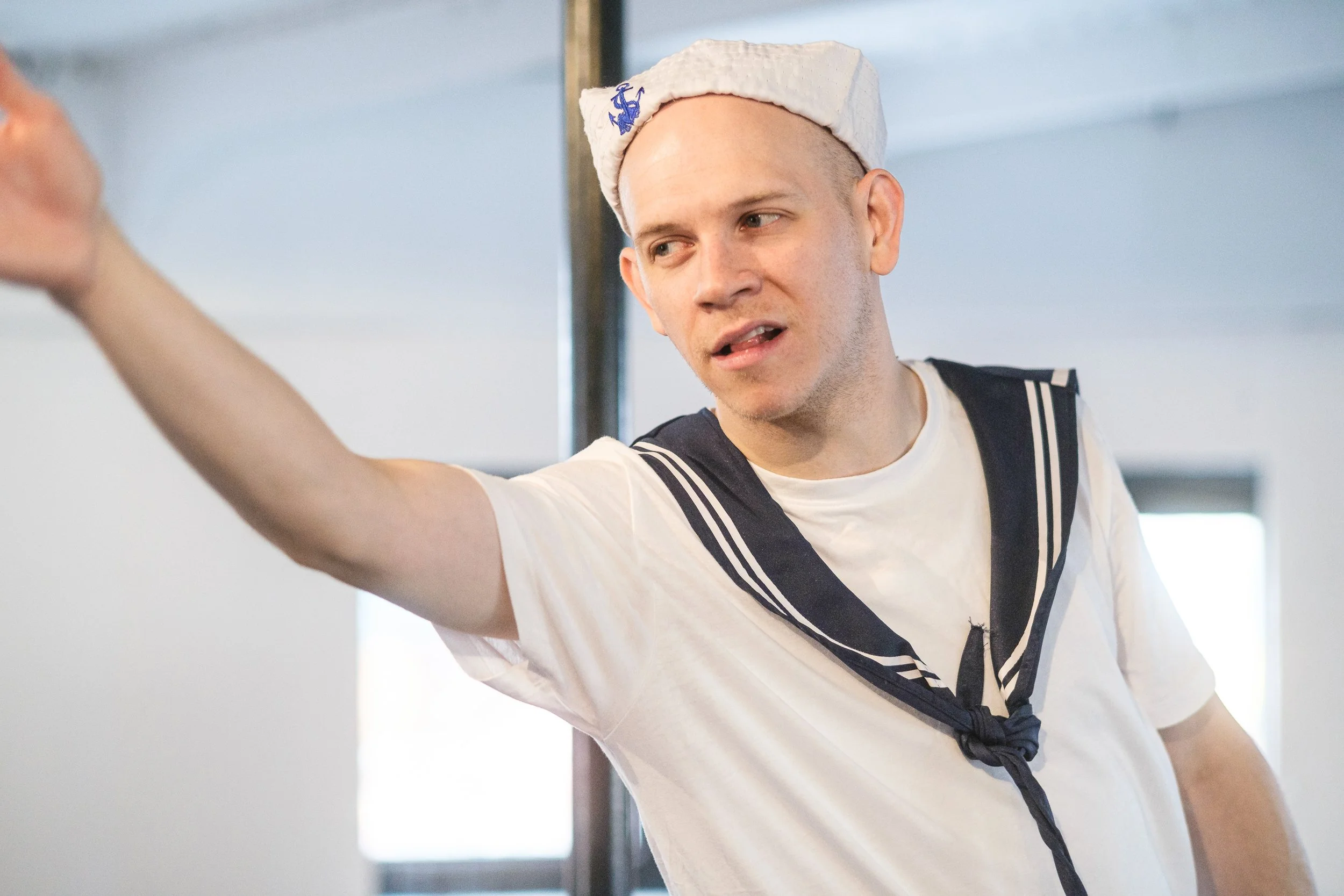 Young man in sailor shirt with sailor hat pointing forward, indoors with a blurred background.