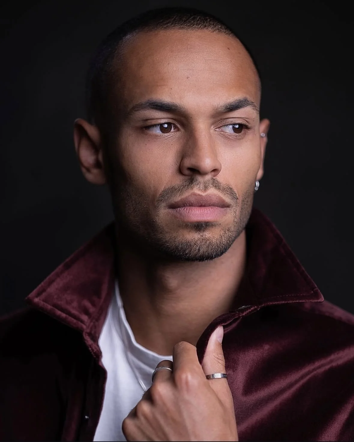 A man with short hair and a beard wearing a burgundy velvet jacket and a white shirt, touching the collar with his right hand, against a black background.