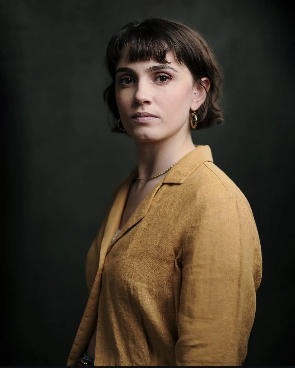 A portrait of a young woman with short brown hair, wearing a mustard yellow blazer, gold earrings, and a gold necklace, against a dark background.
