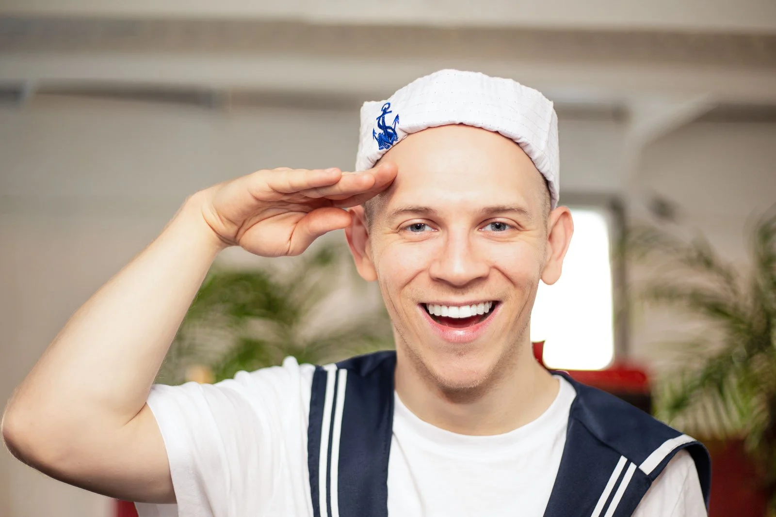 Young man smiling in a sailor uniform saluting, wearing a white sailor cap with an anchor emblem, indoors with a blurred background.