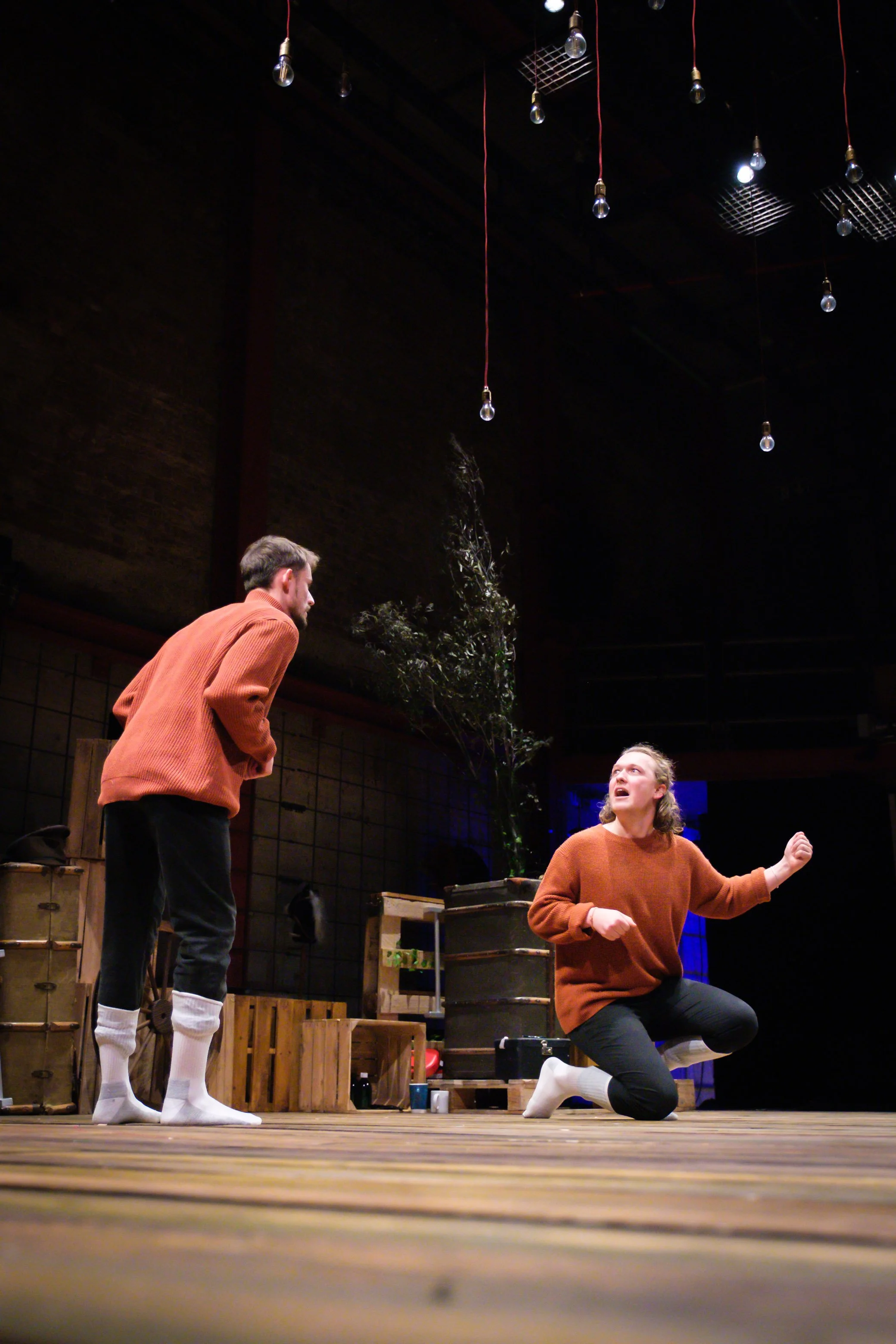 Three people interact on a wooden stage decorated with crates and a potted plant, under hanging light bulbs and a dark ceiling. One person with long hair is kneeling and speaking, while the other two, standing, listen attentively.