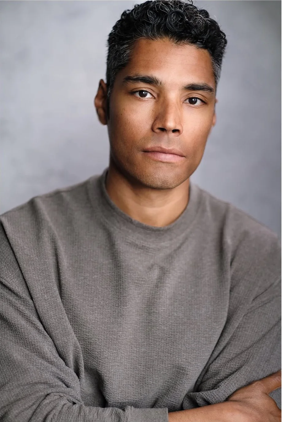 A close-up portrait of a man with short, curly dark hair, wearing a gray long-sleeve sweater, posing with arms crossed against a neutral background.