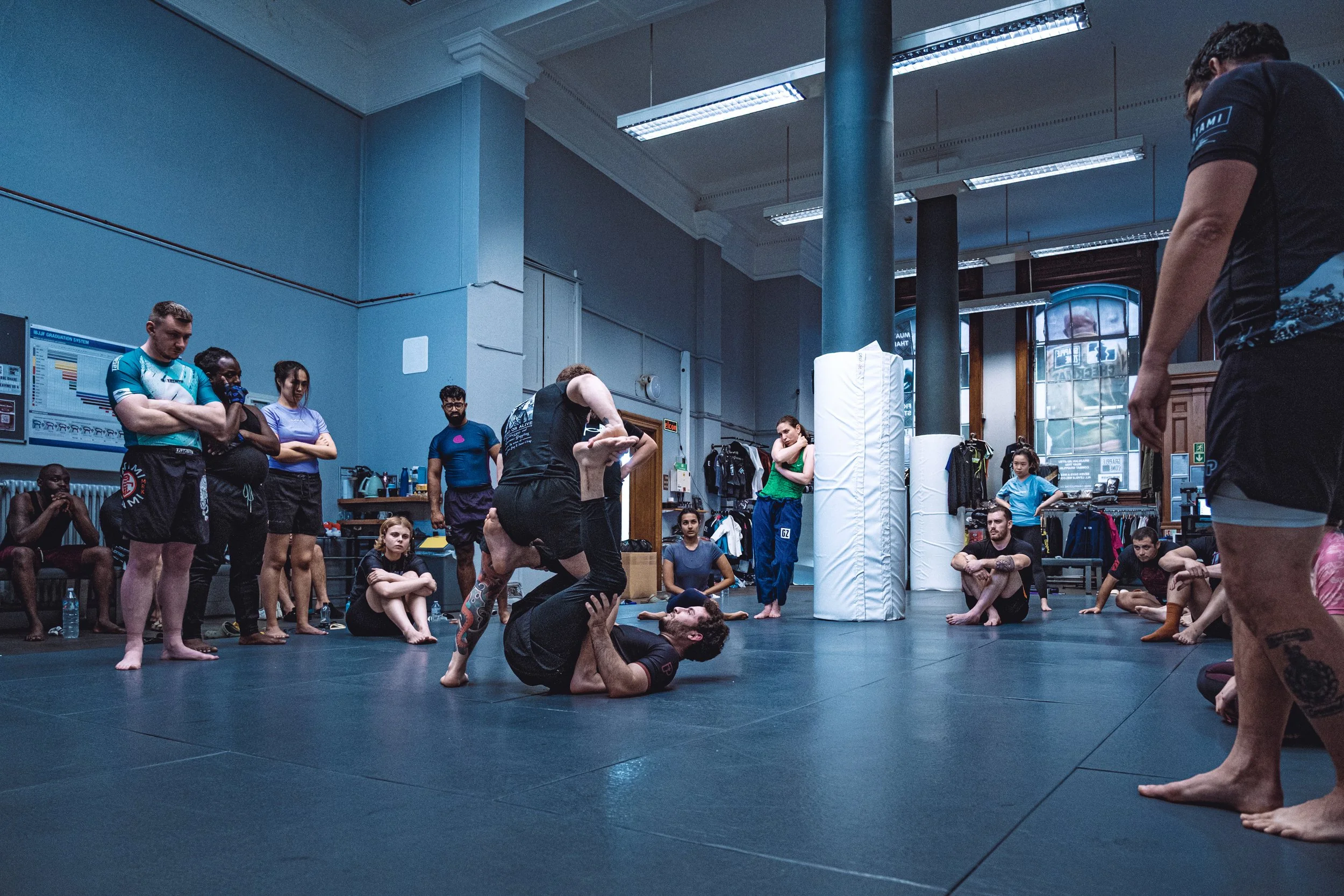 Group of people in a martial arts gym, practicing capoeira, with some standing and others sitting or lying on the mat, and two people in the center engaged in a demonstration. Fight Gym photography. Sports photography.