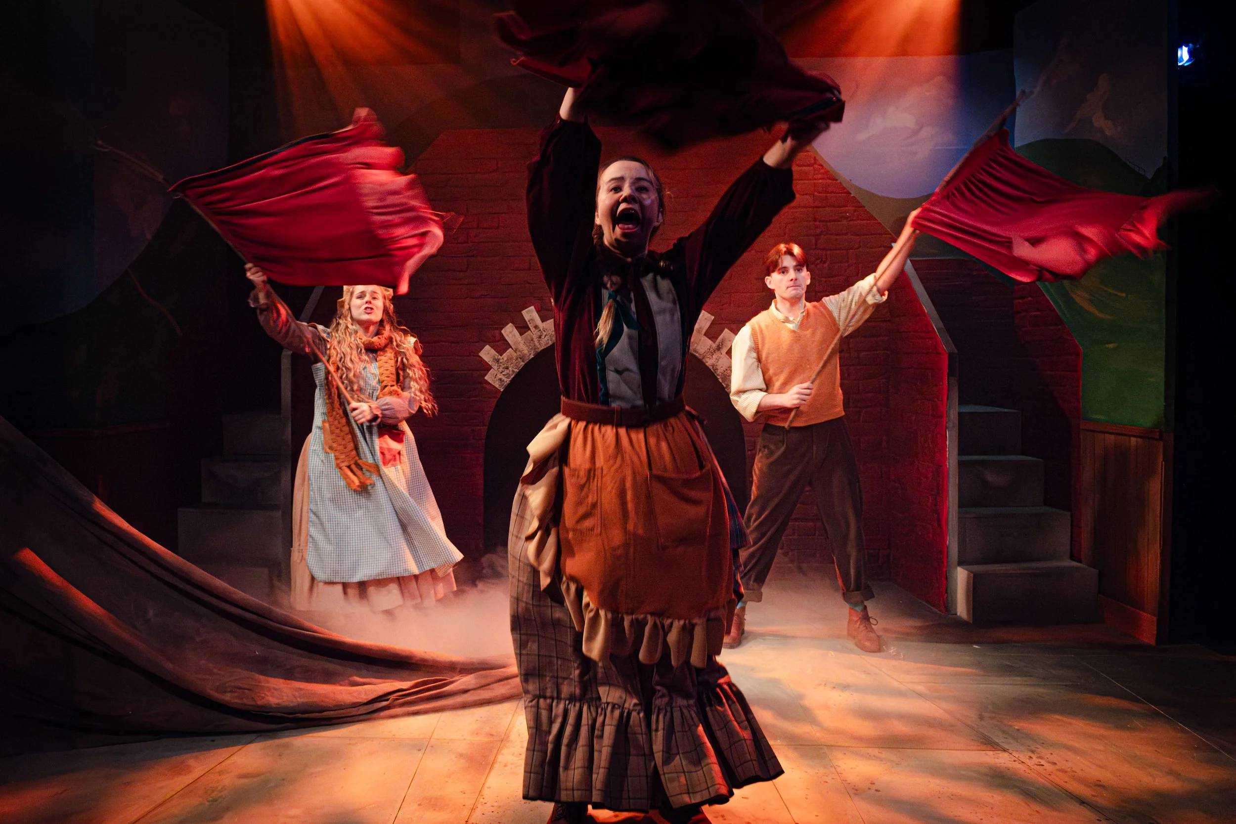 Actors performing on stage in period costumes, holding red flags, with a painted backdrop of a mountain landscape, under warm stage lighting.