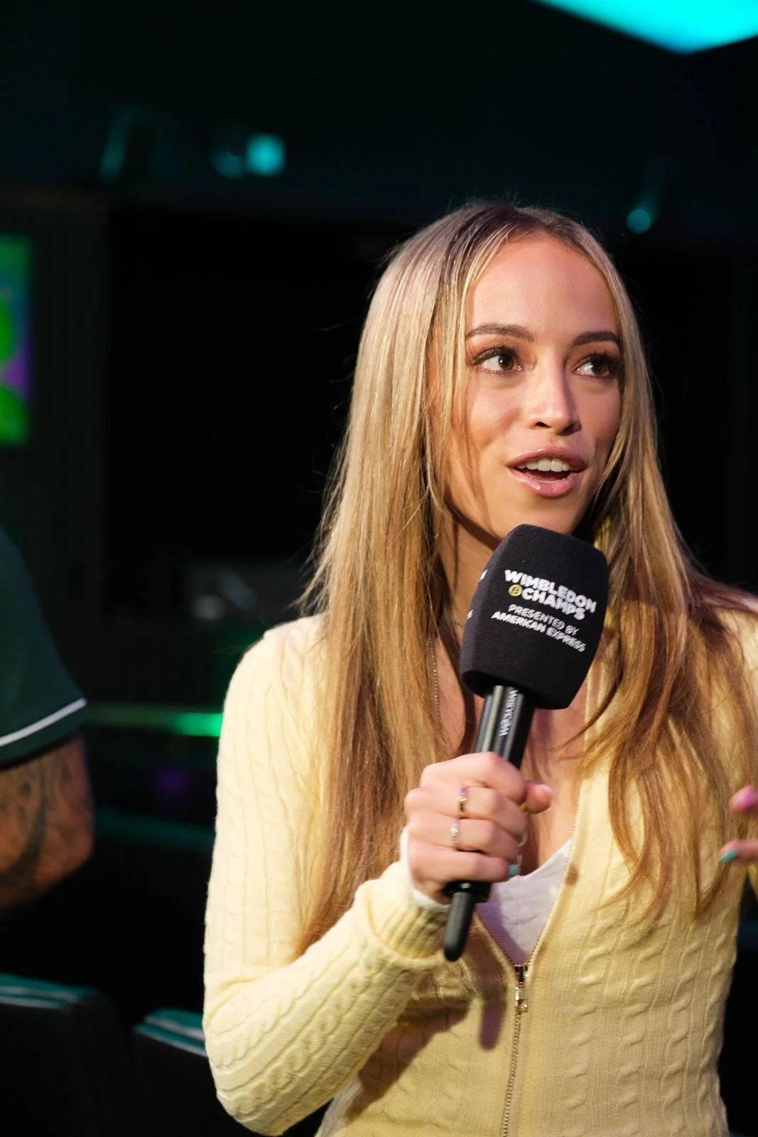 A woman with long blonde hair holding a black microphone with the label 'Wimbledon Champions.' She is wearing a light yellow knit sweater and a white shirt underneath, speaking in an indoor setting.