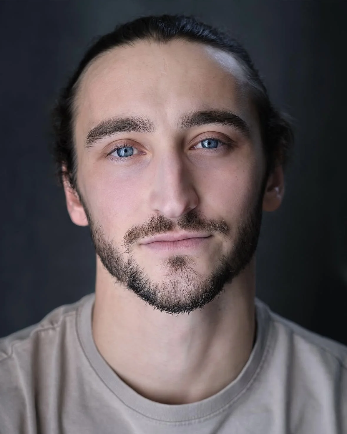 Close-up portrait of a young man with blue eyes, dark hair tied back, and a beard, wearing a light-colored shirt against a dark background.