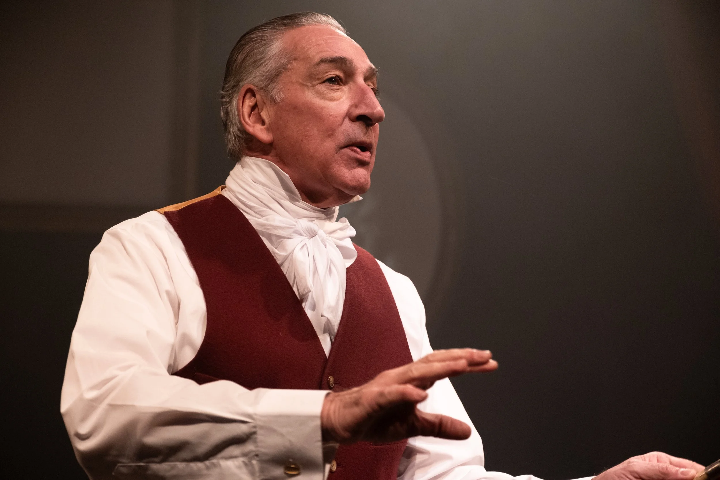 A man dressed as a colonial figure, wearing a white shirt, red vest, and white cravat, standing with hands in front of him.