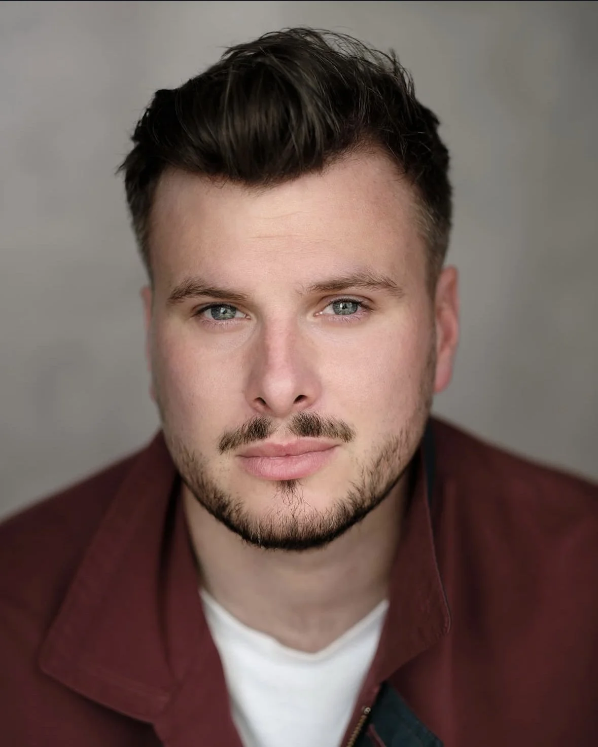 Close-up portrait of a young man with light skin, short brown hair, blue eyes, facial hair, wearing a maroon jacket over a white shirt, against a neutral background.