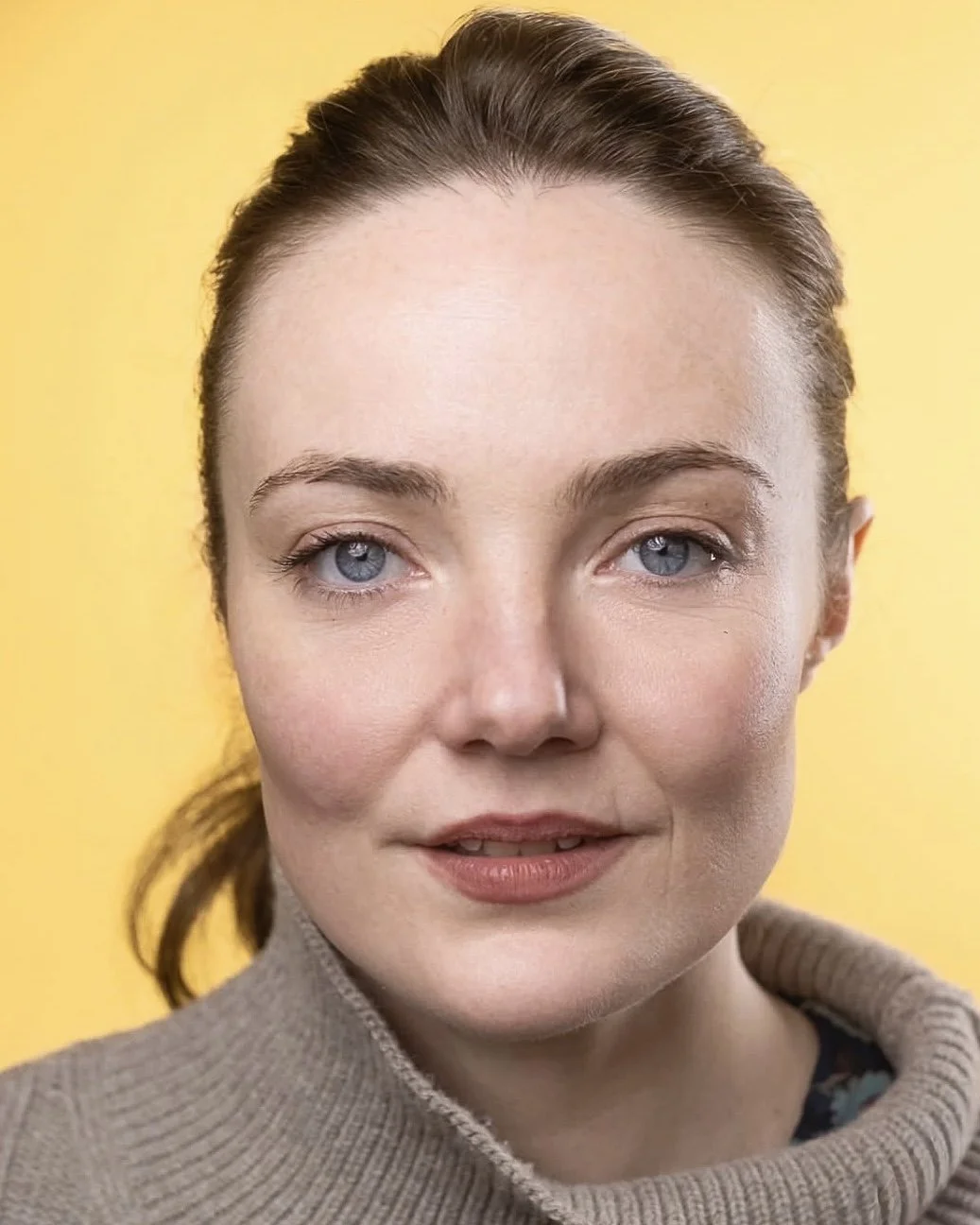 Close-up of a woman with fair skin, blue eyes, and brown hair pulled back, wearing a beige turtleneck sweater, against a yellow background.