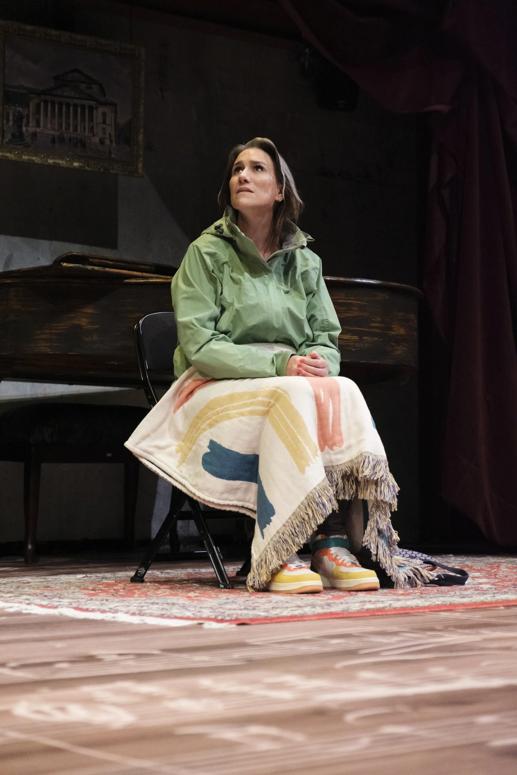A woman in a green jacket sits on a chair on a stage with a colorful blanket over her lap. She looks upward with a thoughtful expression. Behind her, there is a piano, a hanging painting of a building, and red velvet curtains on dark walls. The stage