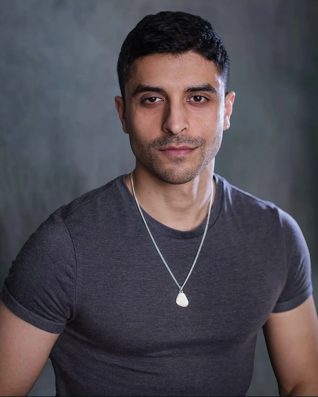 A young man with short dark hair and a slight beard, wearing a dark grey t-shirt and a silver necklace with a large white pendant, looking directly at the camera with a neutral expression against a grey background.