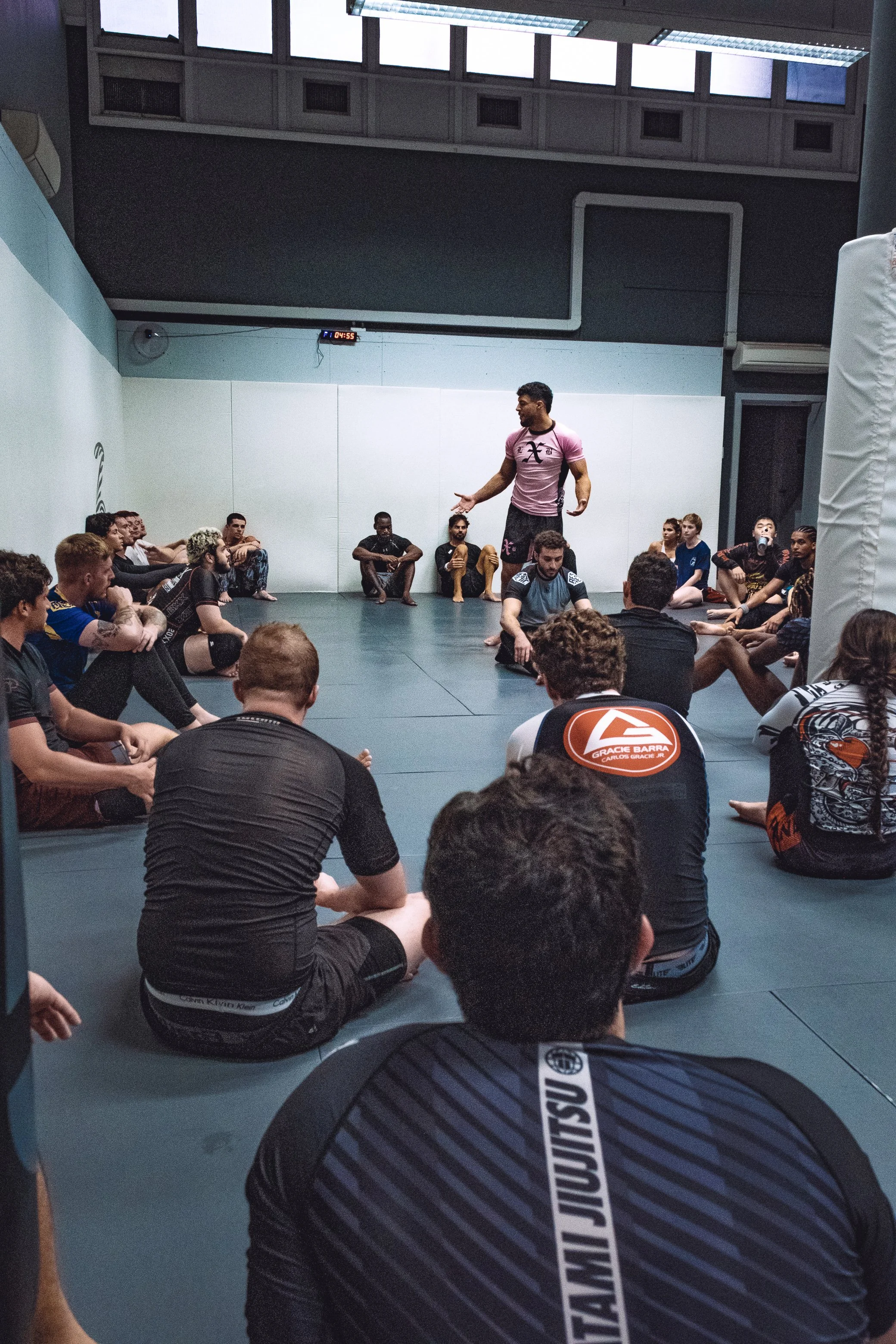 A group of martial arts students sitting on the mat in a gym listening to their instructor, who is standing and speaking in front of them. The instructor is wearing a pink shirt, and the students are wearing black and colorful uniforms.