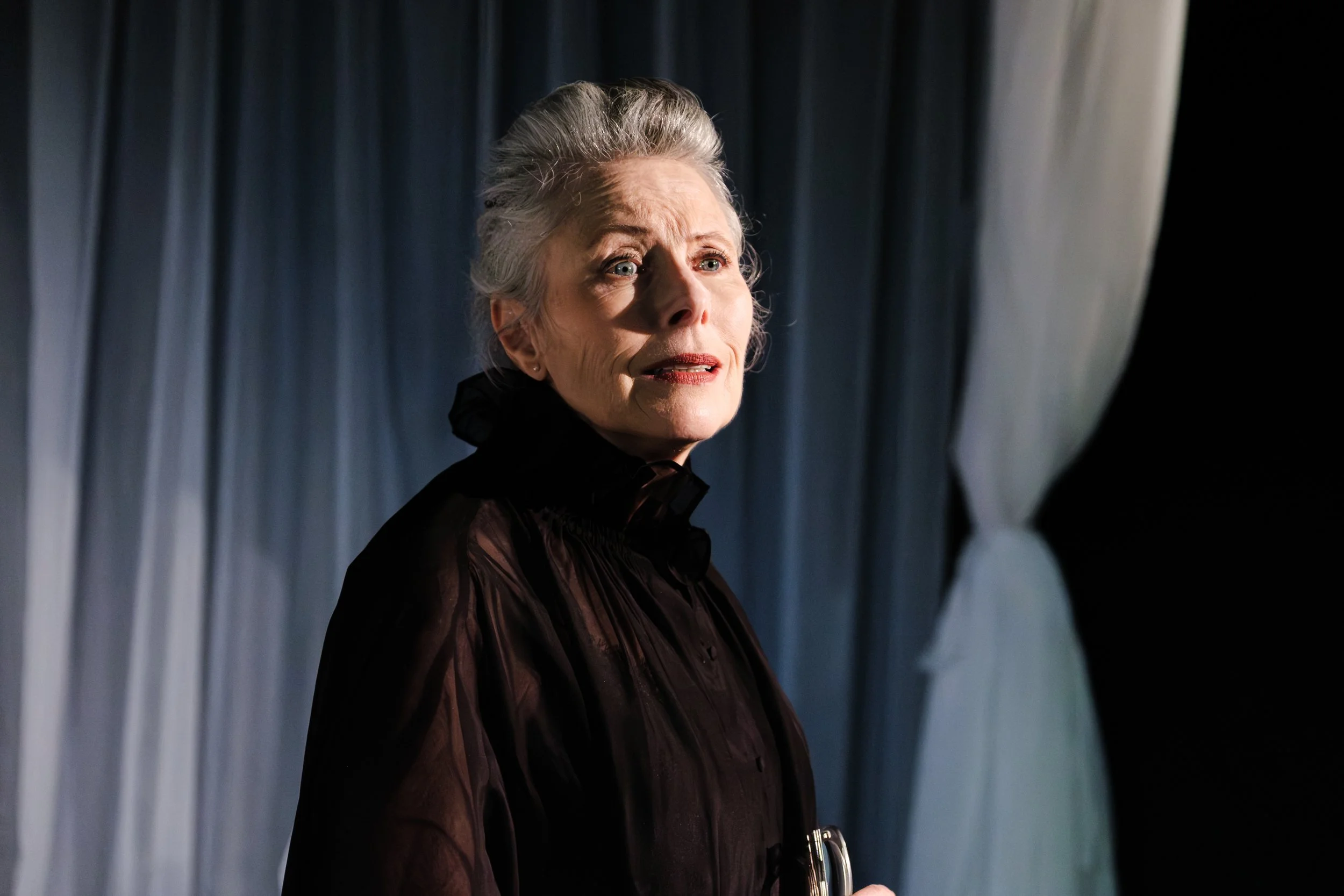 A woman with gray hair styled in an updo, wearing a black dress, looking to her left with a serious expression, standing in front of dark curtains.