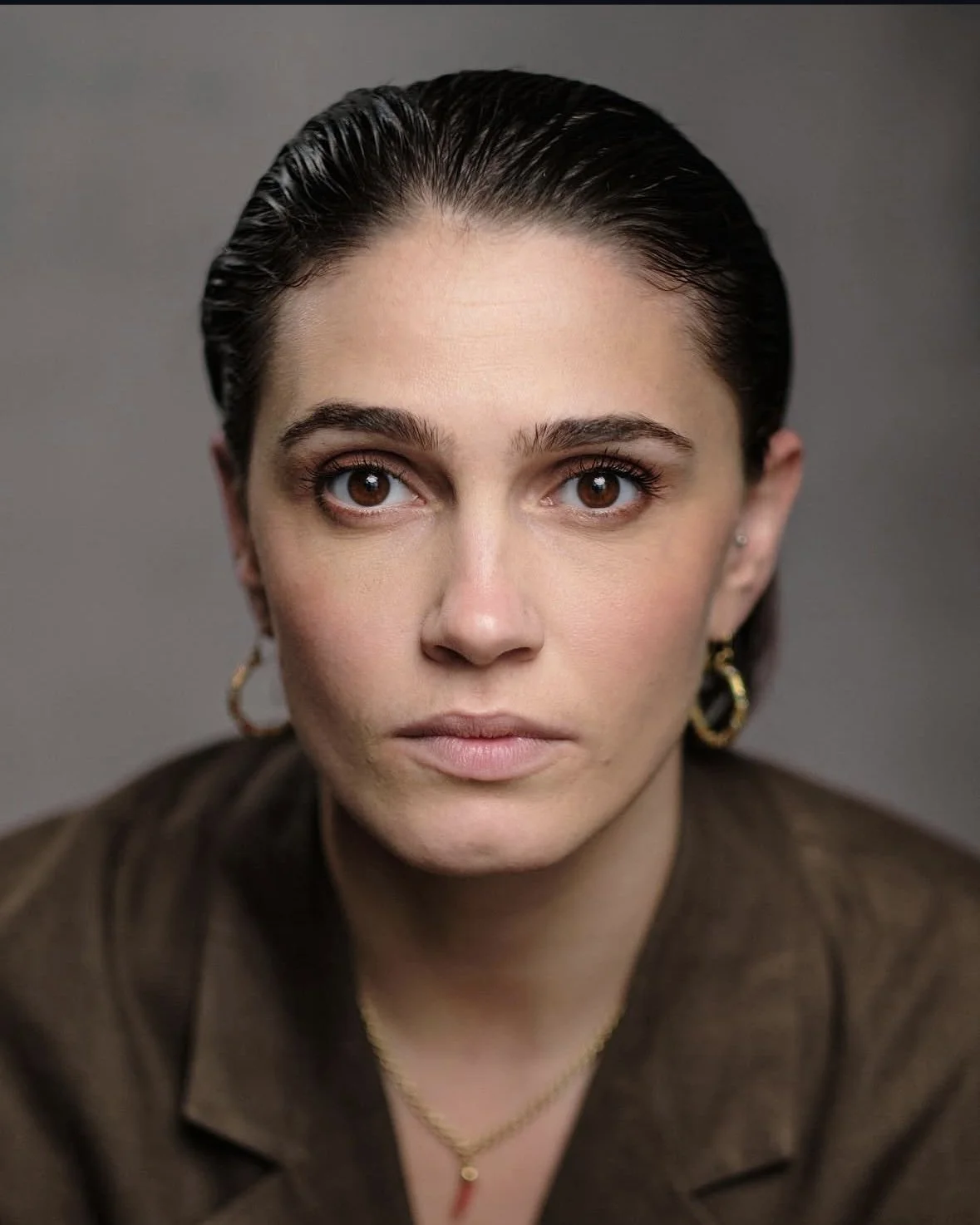 A woman with dark hair slicked back, wearing gold earrings and a gold necklace, looking directly at the camera with a neutral expression.