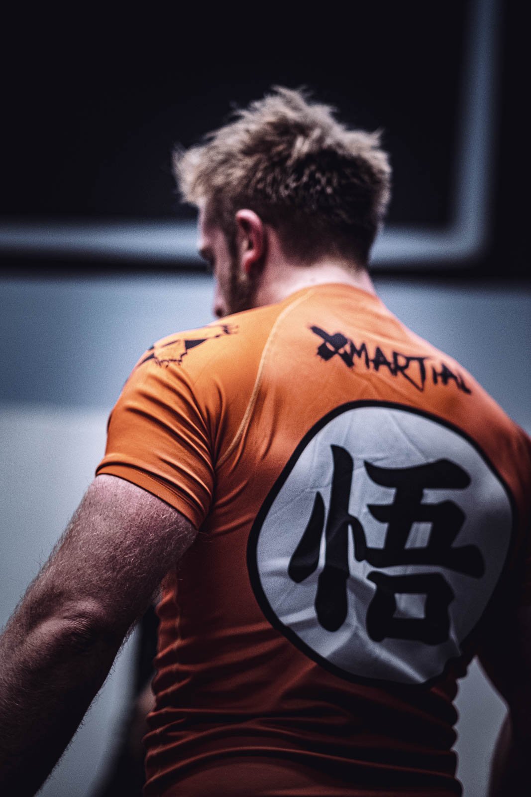 A person with spiky, blonde hair wearing an orange sports shirt with a white circle emblem containing a Japanese character on the back of the shirt. Grappling bjj sports photography