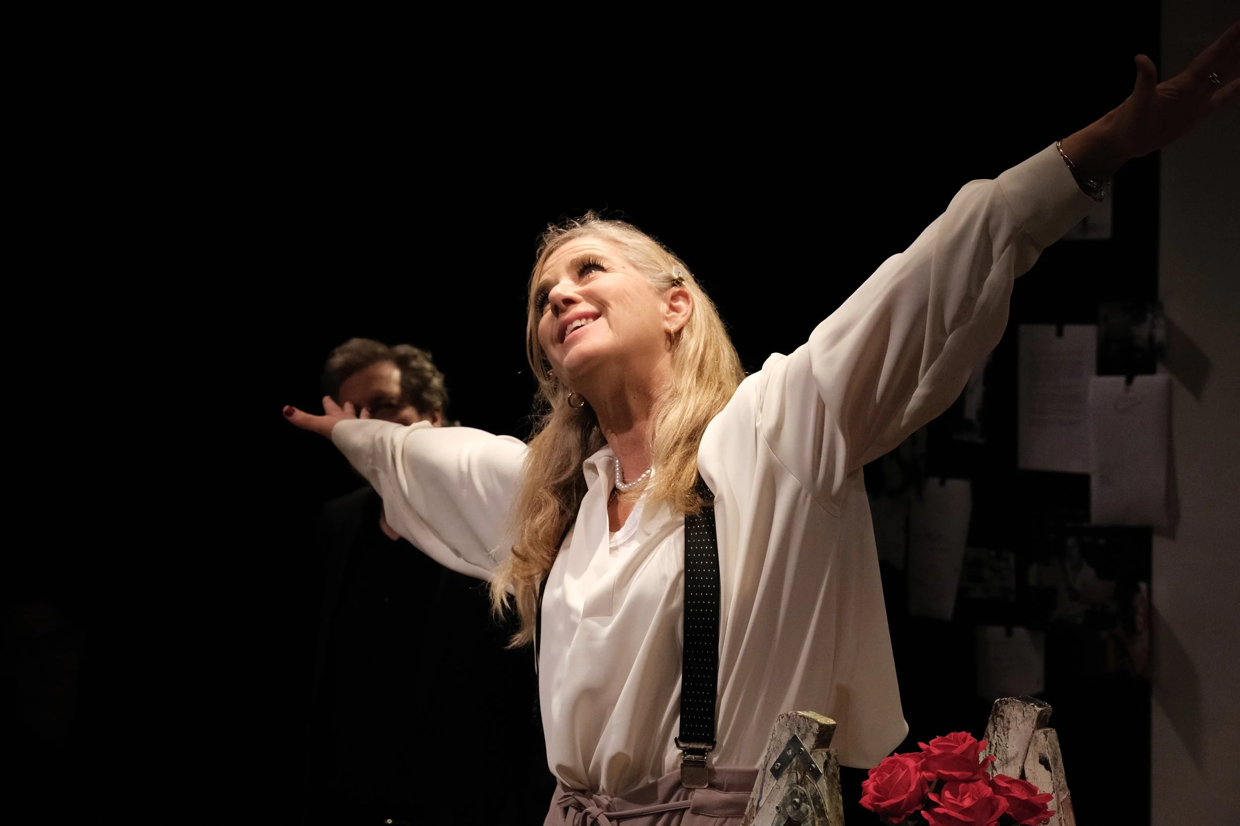 An older woman with blonde hair, wearing a white blouse, smiling with arms outstretched, standing in front of a dark background with people and red roses in the foreground.