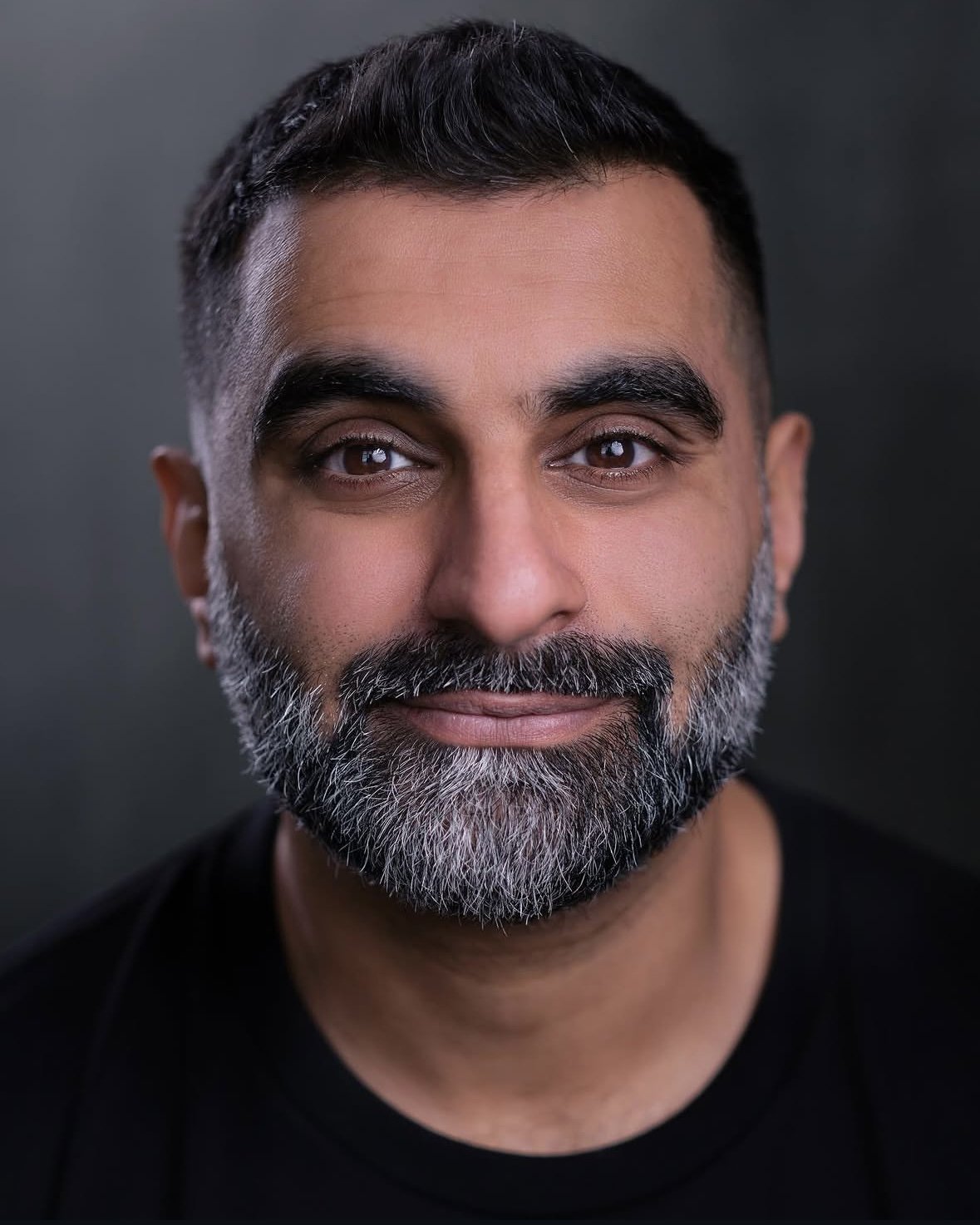 Close-up portrait of a man with dark hair, brown eyes, and a salt-and-pepper beard, wearing a black shirt against a dark background.