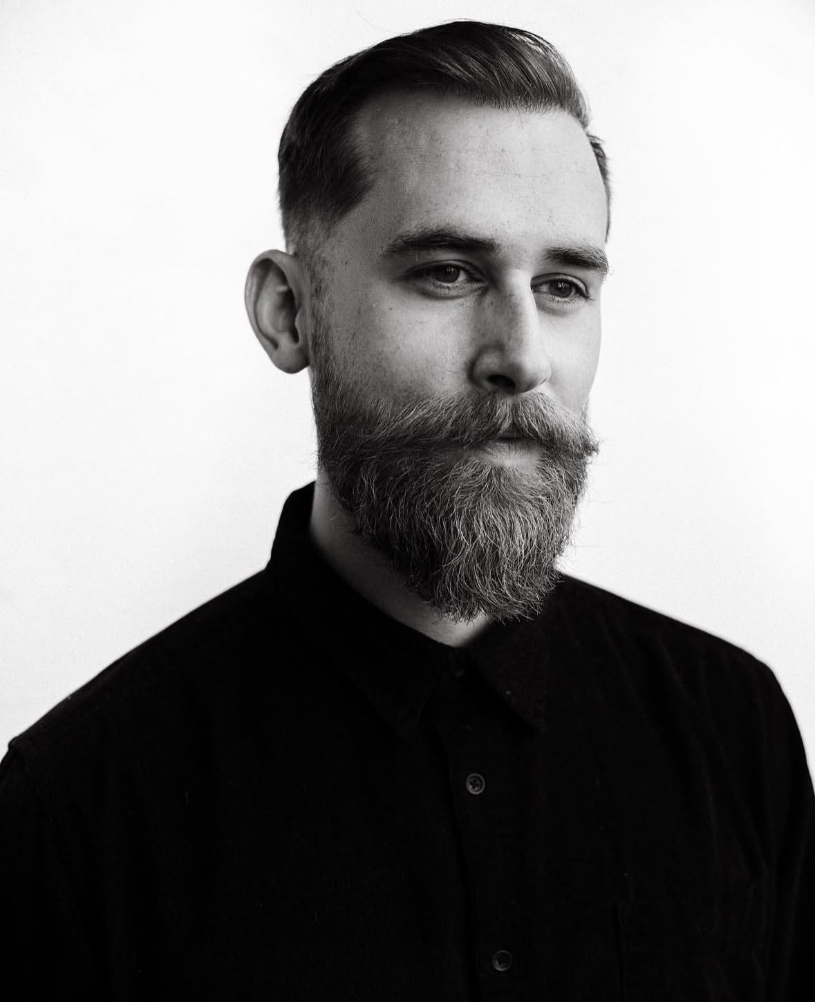 Black and white portrait of a man with a trimmed hairstyle and a full beard, wearing a dark shirt, against a plain background.