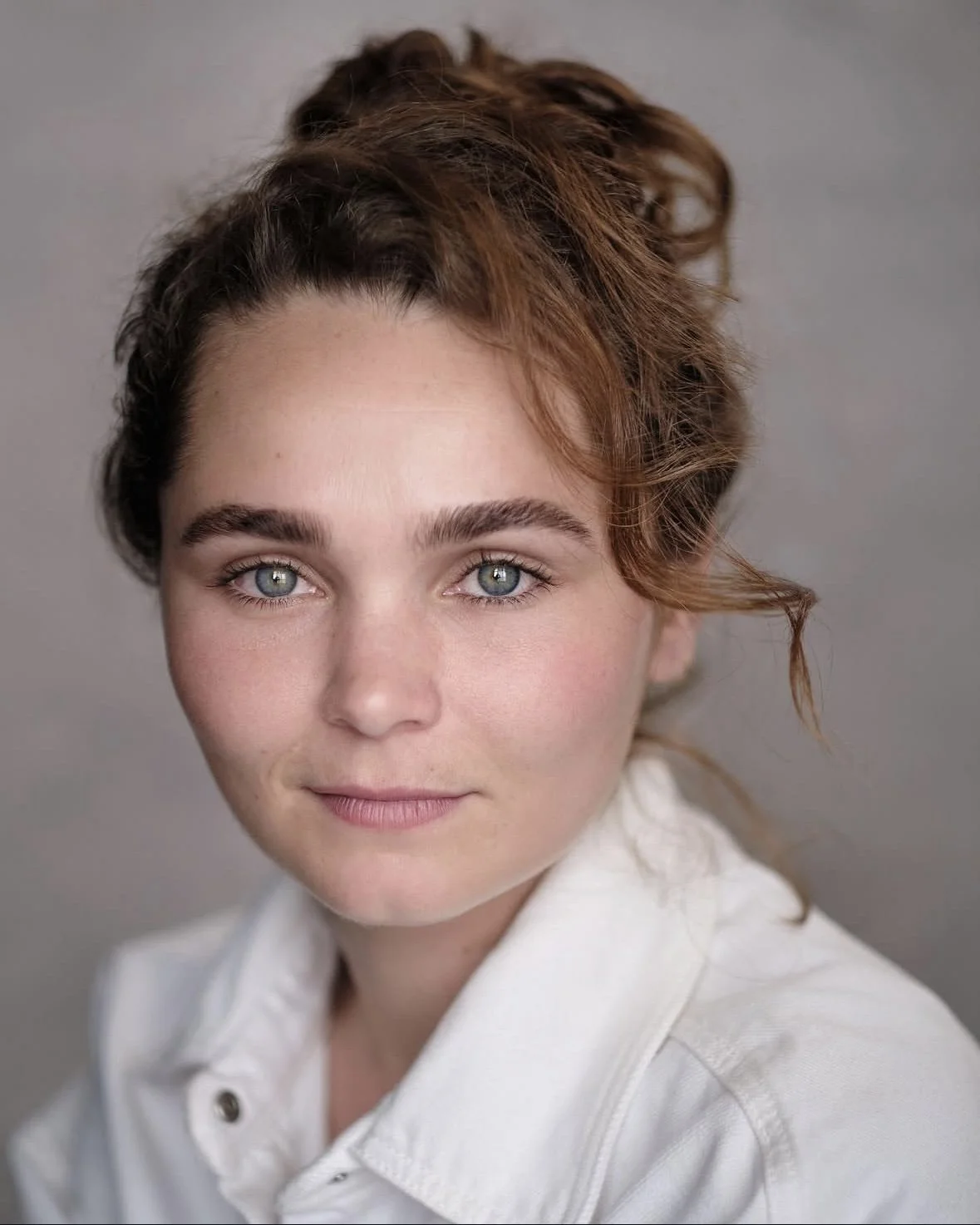 Close-up portrait of a woman with blue eyes, red wavy hair, and a slight smile, wearing a white shirt against a neutral background.