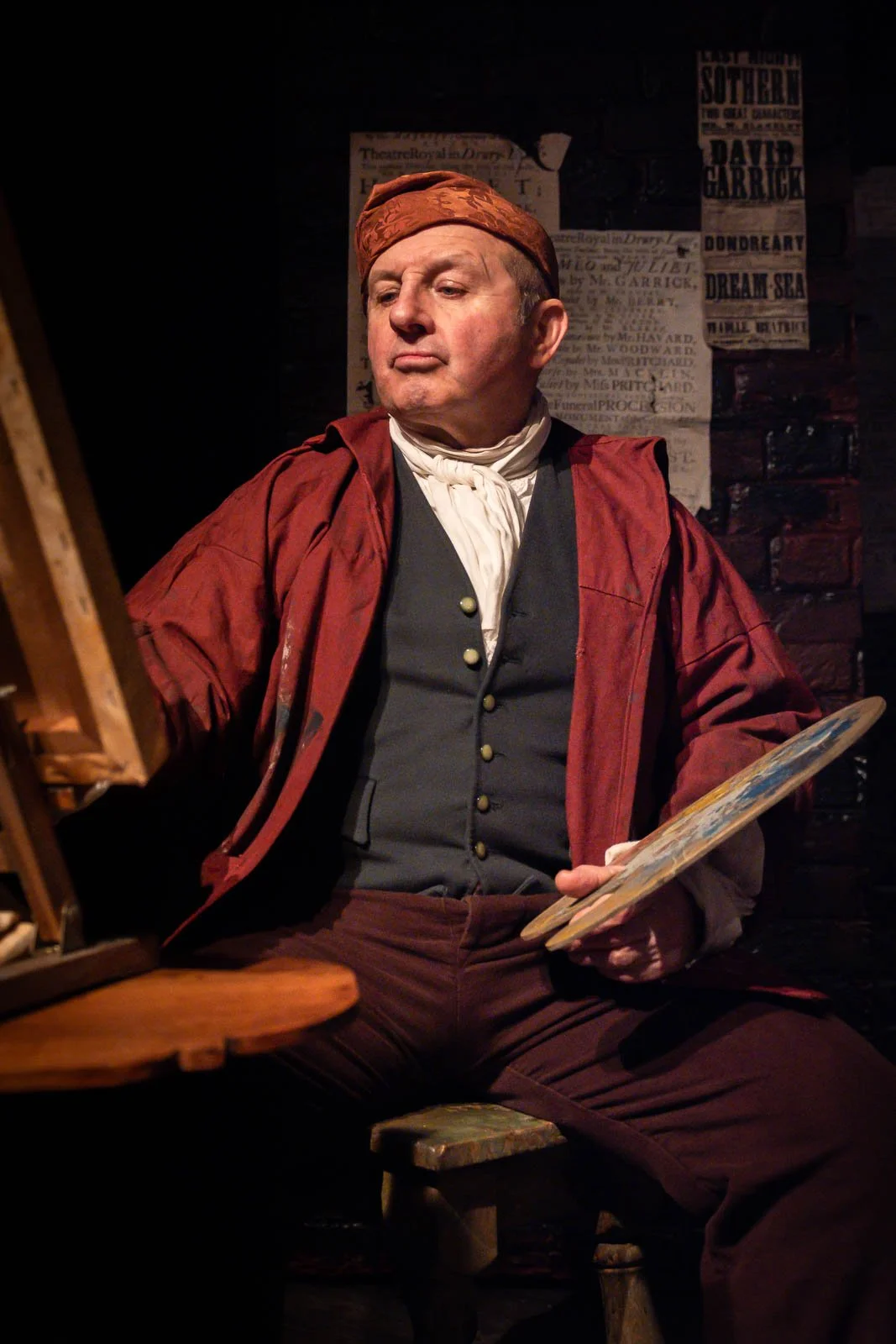 A man dressed in historical clothing, including a red coat and a brown cap, sitting in a dimly lit room with old posters on brick wall behind him, holding a painter's palette. Theatre Photography. Theatre show. Theatre photo. Performance Photo
