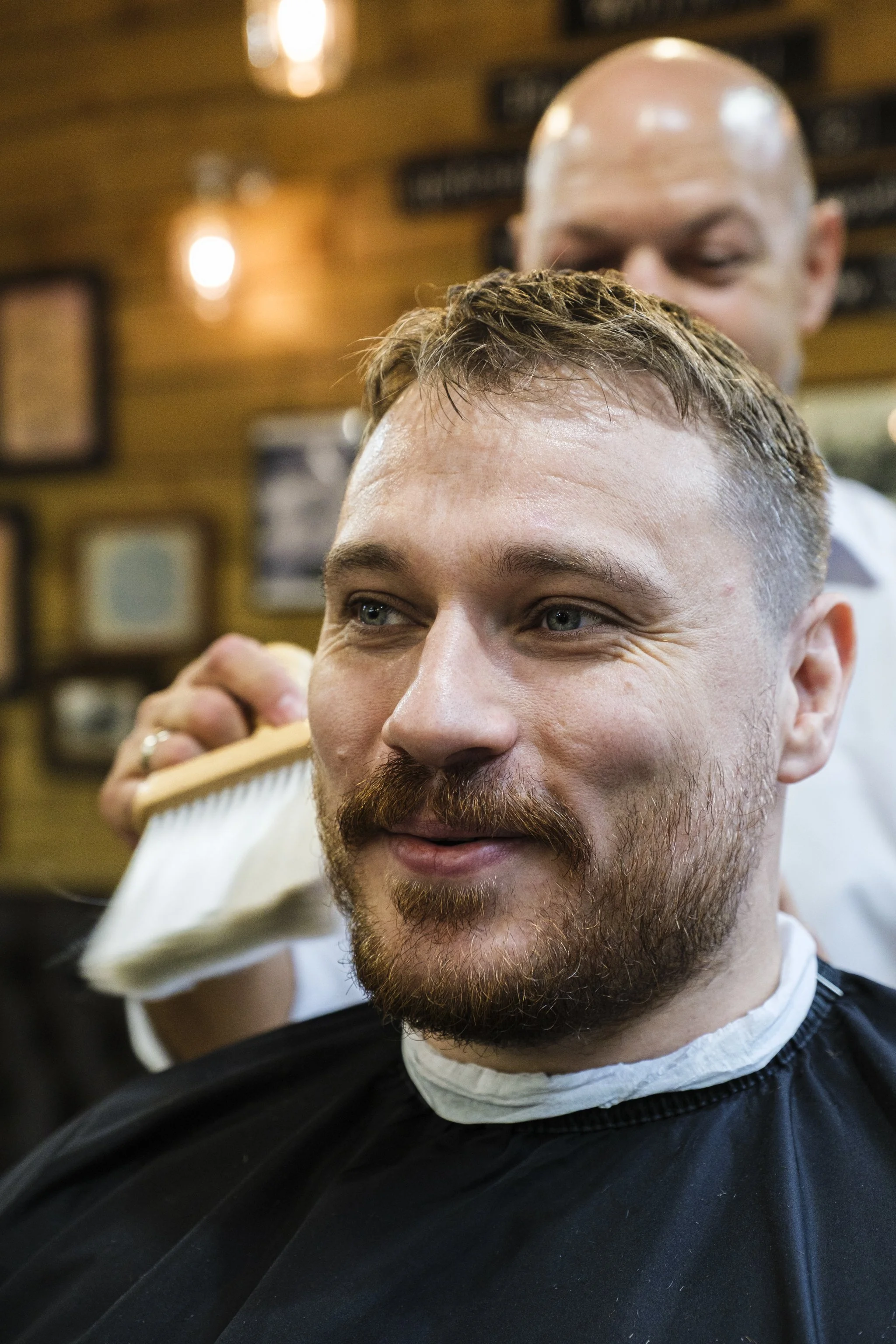 A man with a beard and mustache getting a haircut in a barbershop, with a barber standing behind him, wearing a white shirt. The background has warm lighting and framed pictures on the wall.