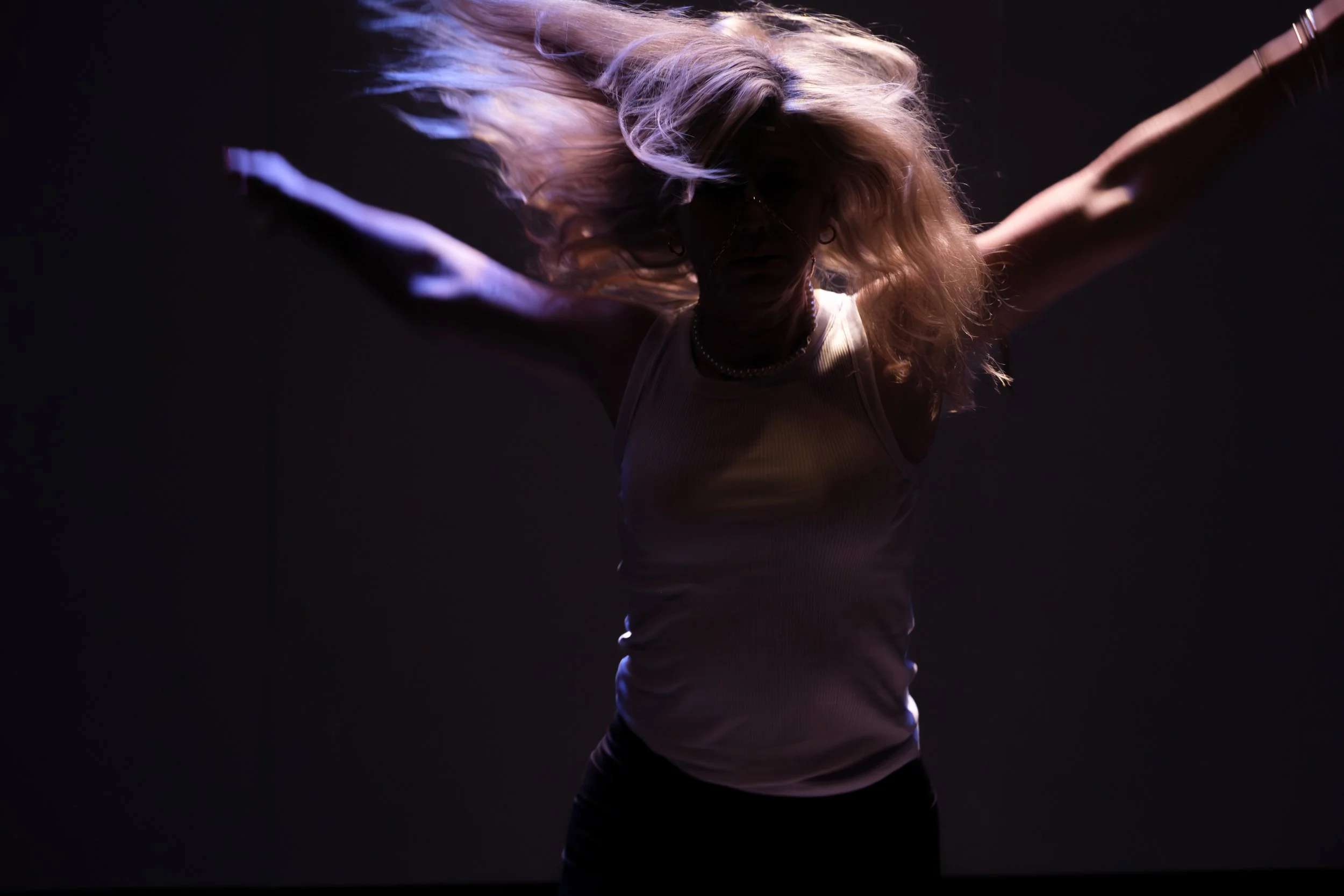 A woman with long, wavy blonde hair wearing glasses and a white tank top, dancing or moving energetically in a dark environment with subtle purple lighting.