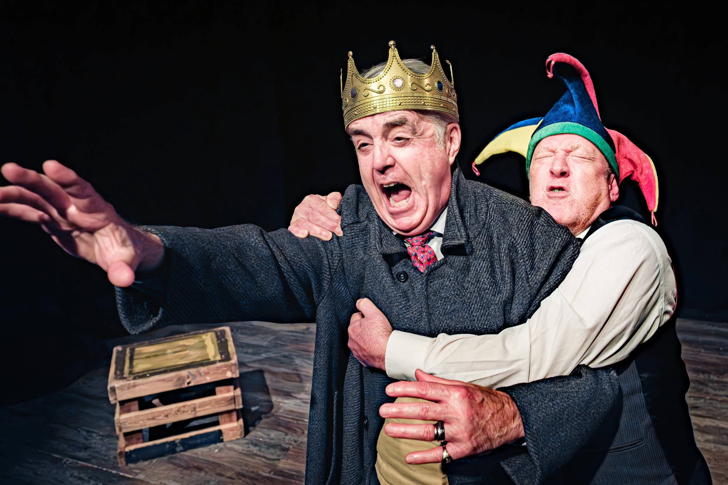 Two men dressed as a king and a jester playfully engaged in dramatic poses; the man as a king wears a gold crown, and the man as a jester wears a colorful, drooping hat, both making expressive facial gestures against a dark background.