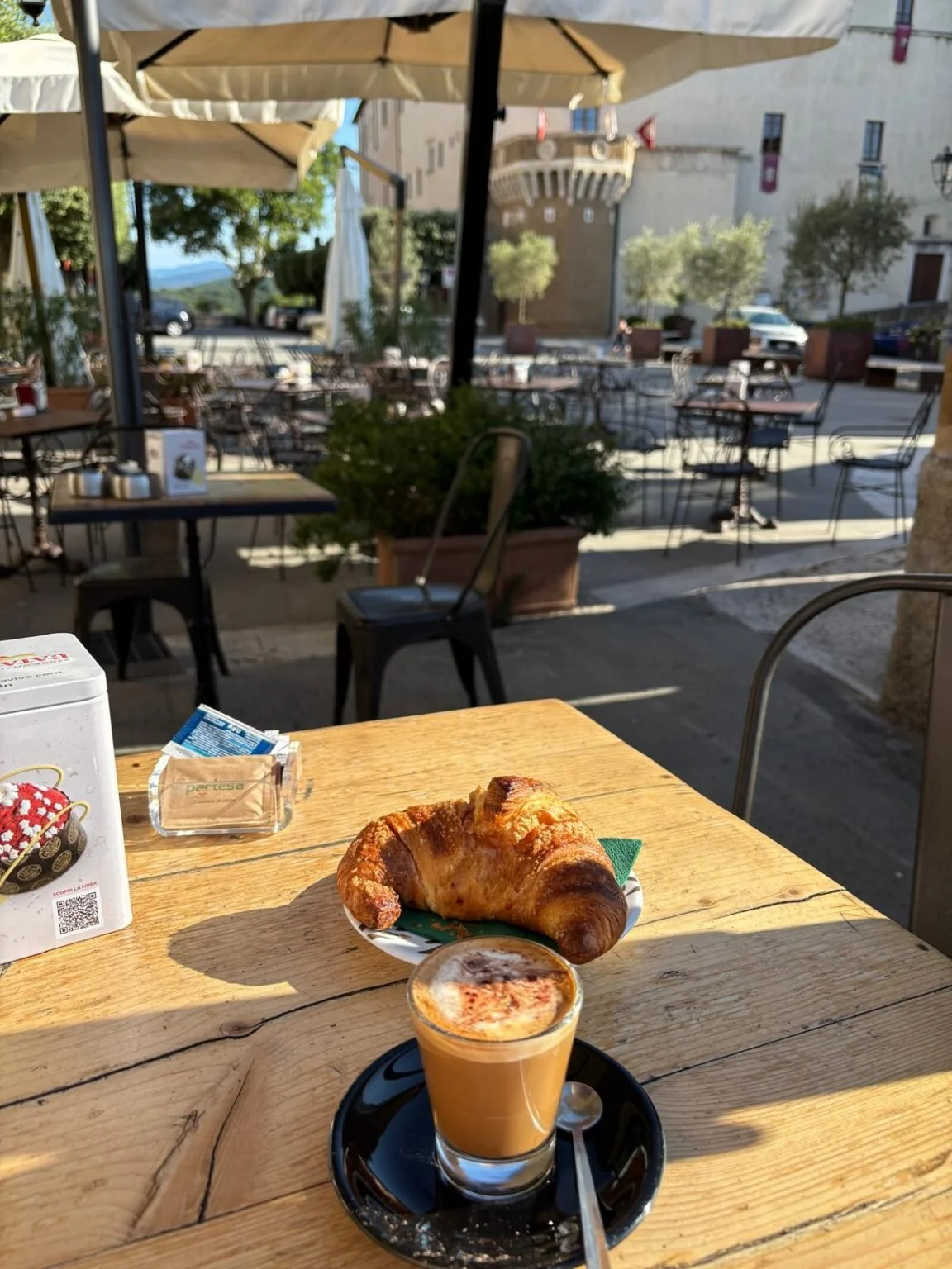 A croissant and a small glass of coffee on a wooden table at an outdoor café with umbrellas, potted plants, and a historic building in the background.