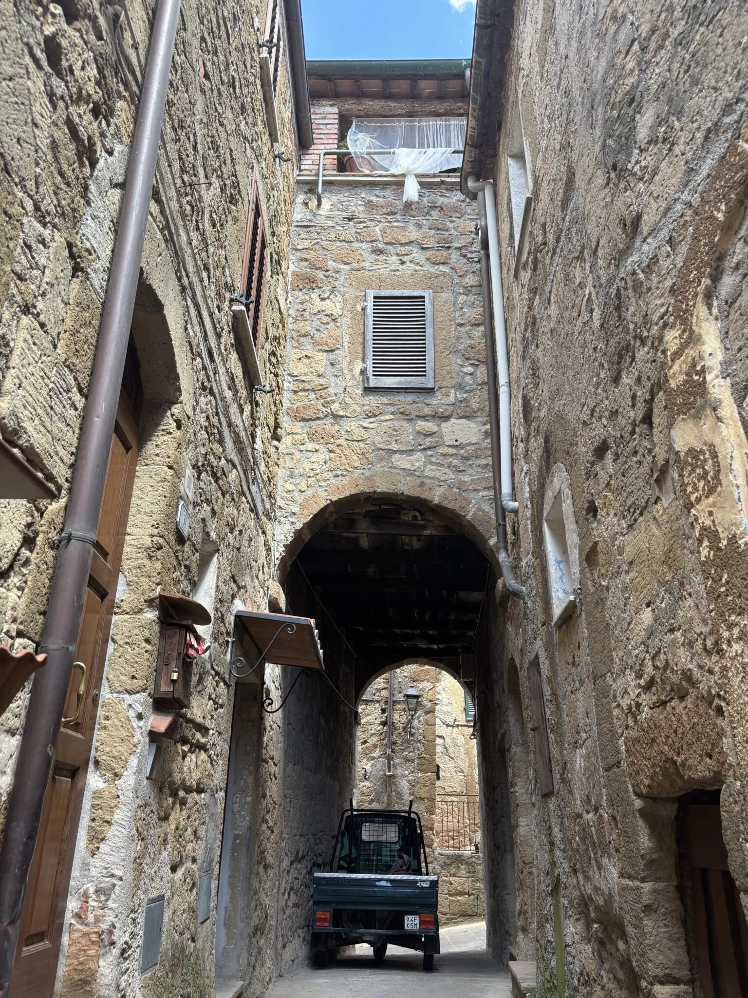 A narrow alleyway between old stone buildings with a small truck parked underneath an archway, small windows with brown shutters, and a hanging clothesline with a white curtain. Some metal pipes run along the walls.
