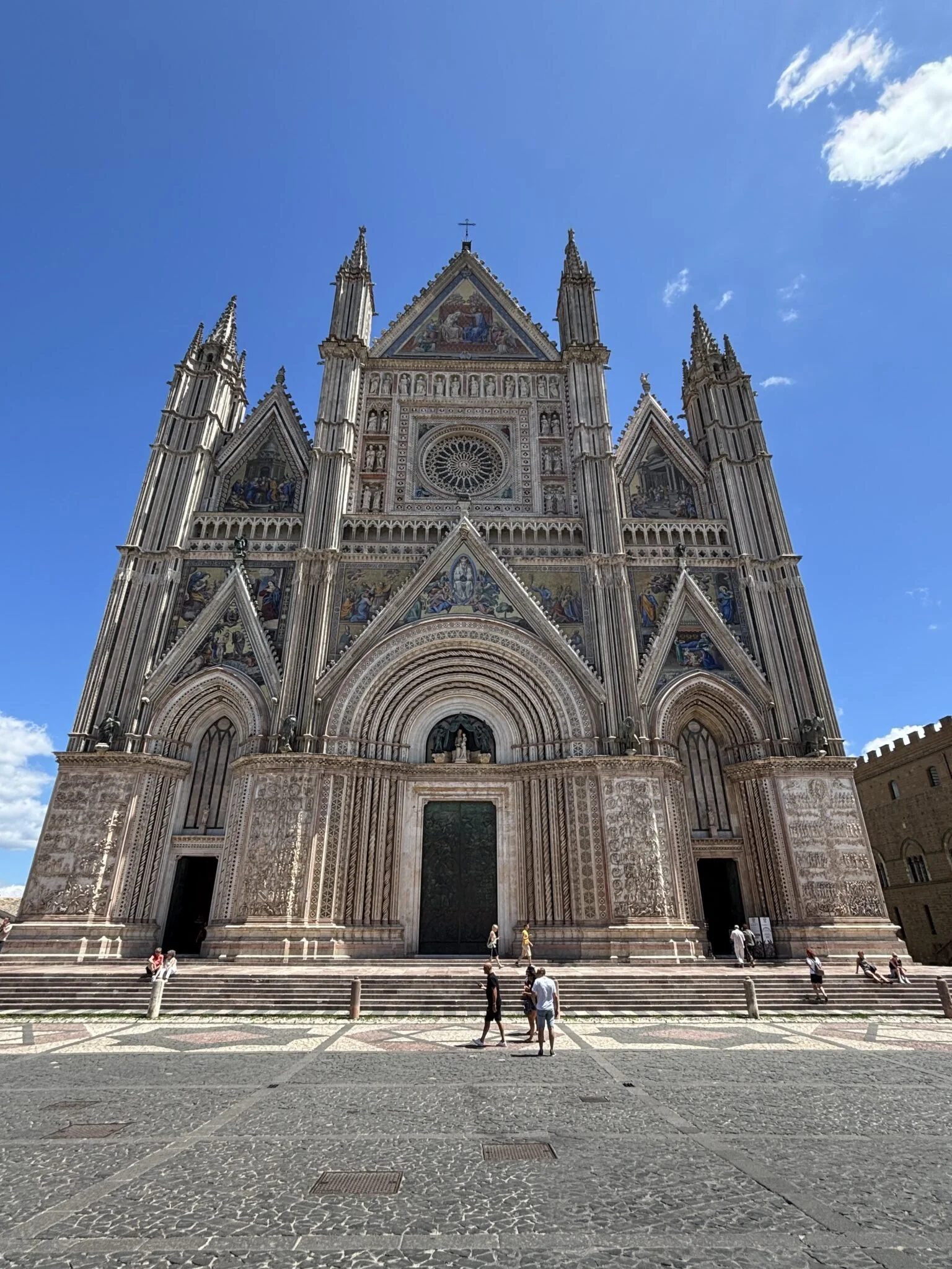 Front view of a grand cathedral with detailed stone carvings and colorful mosaics on the facade, a rose window at the center, and spires reaching into a blue sky. People are walking and sitting on the steps in front.