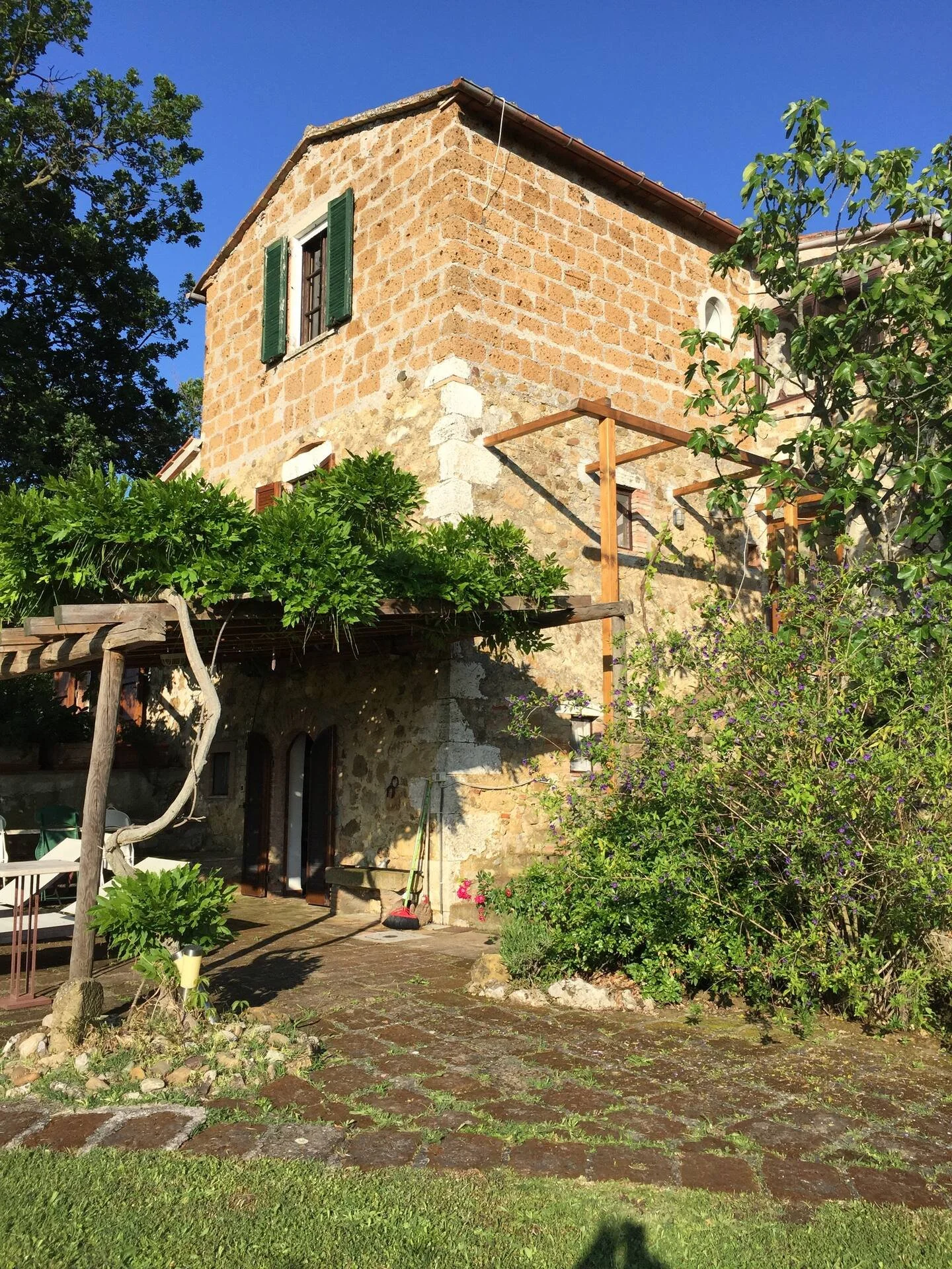 Stone house with green shutters, surrounded by lush greenery and flowering bushes, with a stone patio and a wooden pergola in the backyard.