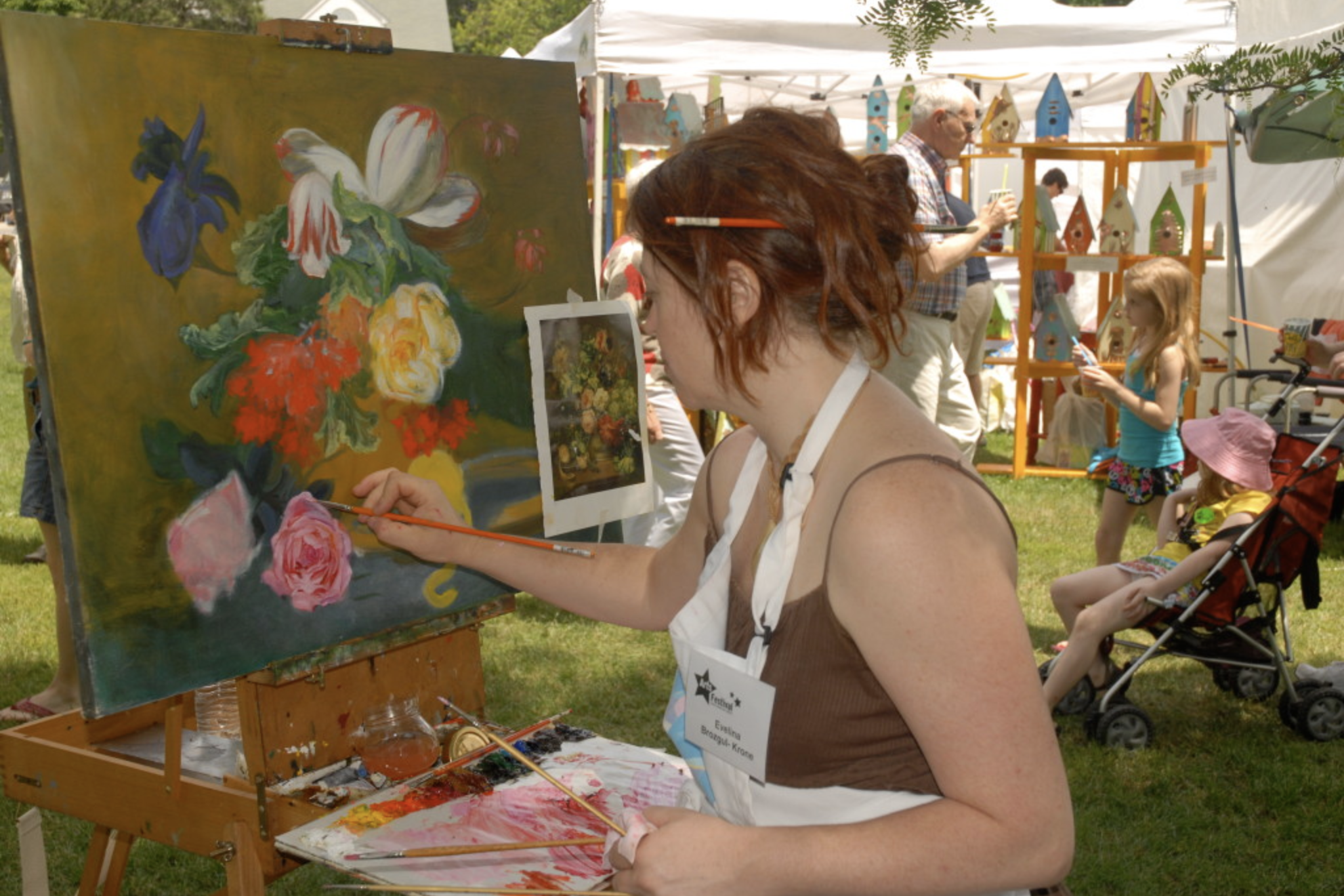 A woman painting a colorful floral still life on a canvas at an outdoor art festival, with people, tents, and birdhouse crafts in the background.