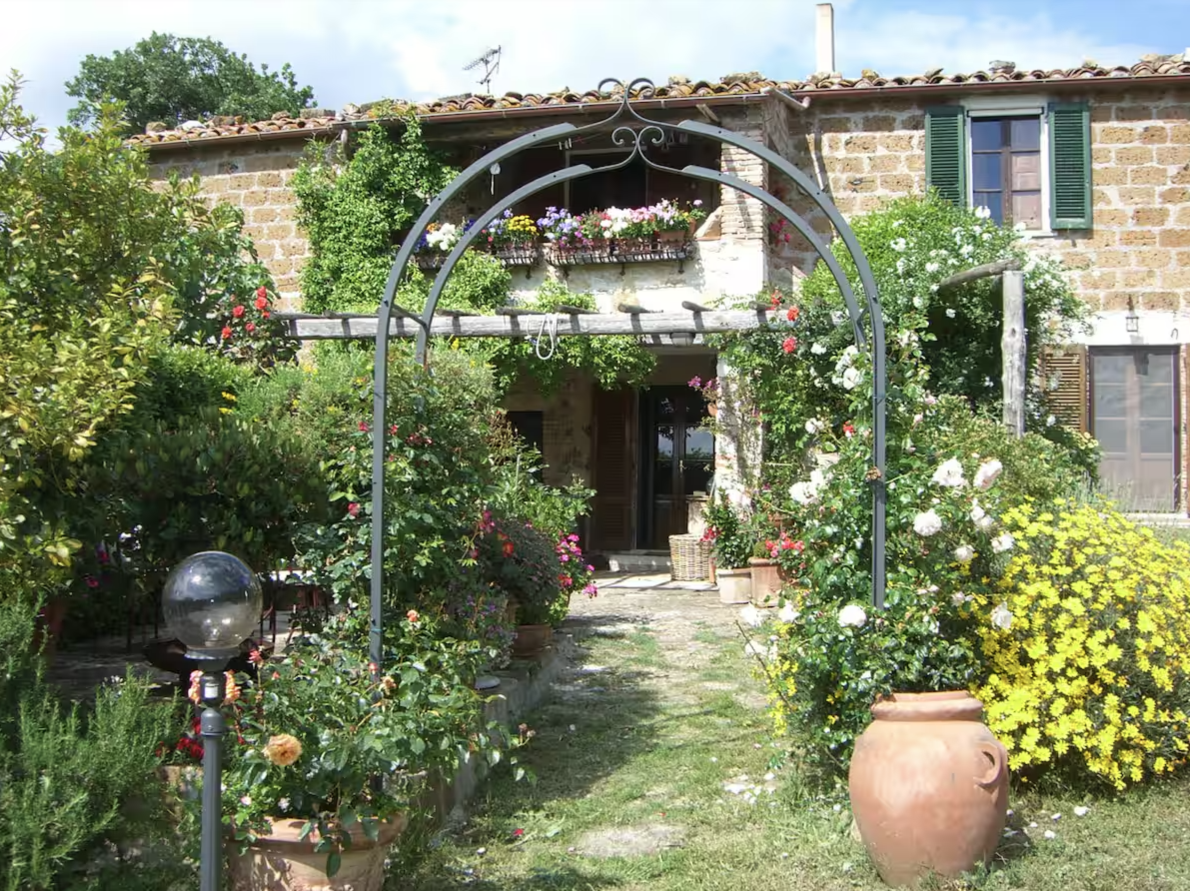 A lush garden with colorful flowers, a stone house with green shutters, and a large terracotta pot in the foreground. There is a metal archway covered with flowers, leading to a patio area.