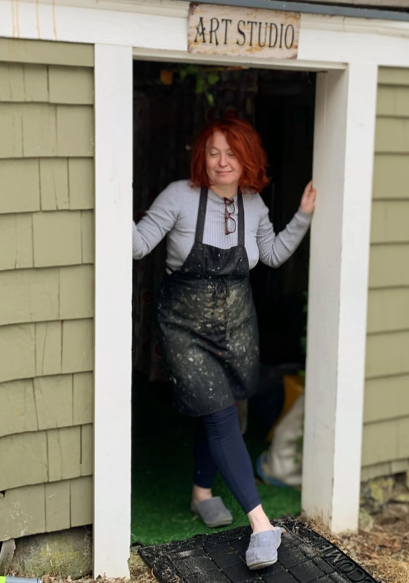 A woman with red hair wearing glasses, a striped shirt, and an apron standing in a small art studio door, smiling and wearing grey house slippers. The studio is labeled 'Art Studio' above the door. The exterior wall is house siding, and the woman is standing on a green artificial turf mat.