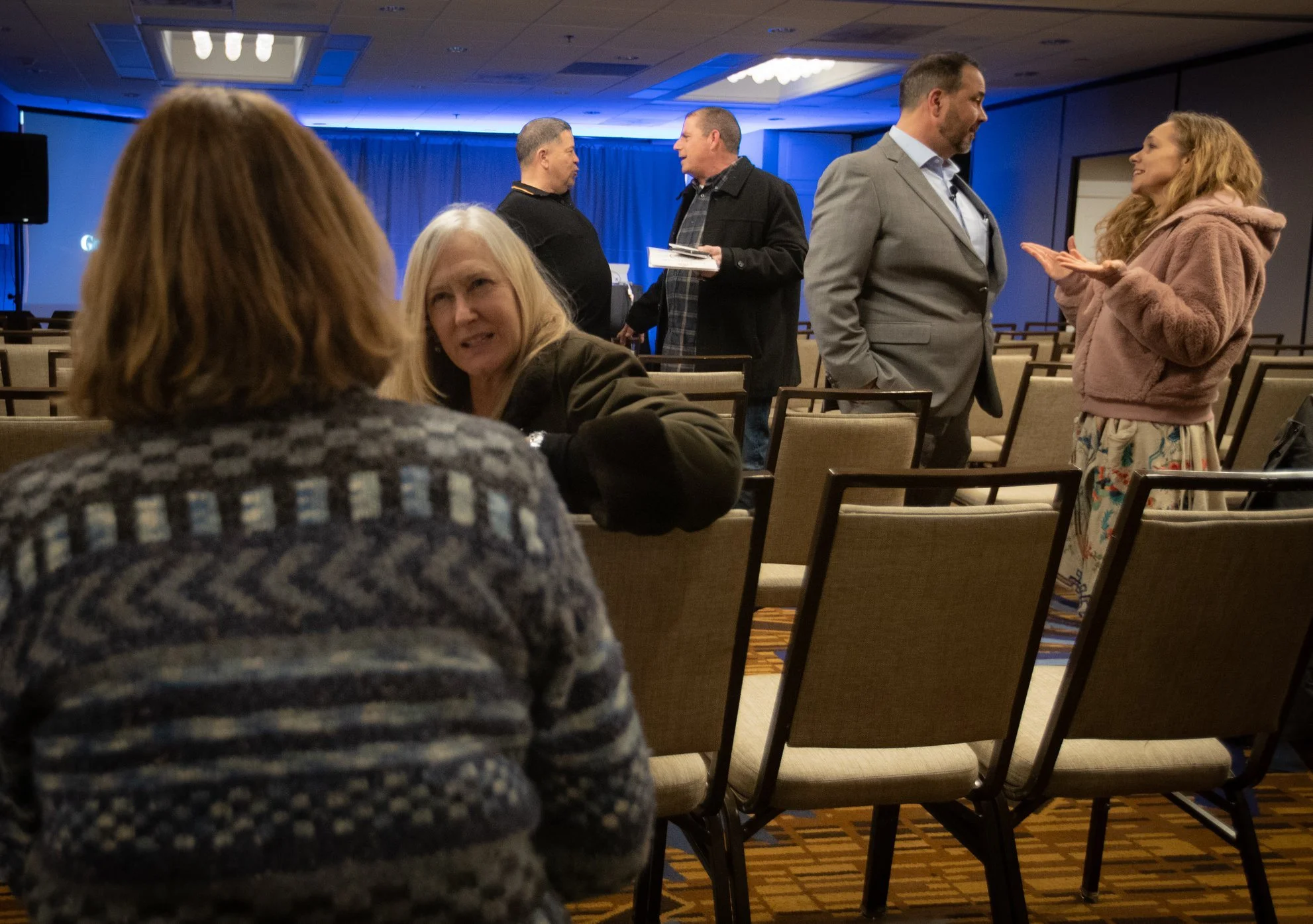 People talking and interacting in a conference or event room with chairs, blue lighting, and a presentation screen in the background.