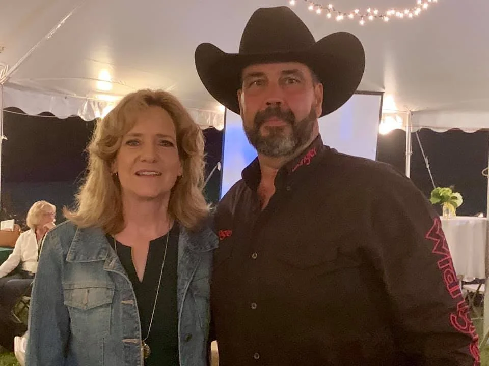 A woman with blonde hair wearing a denim jacket standing next to a man in a cowboy hat and black shirt at an indoor event with string lights.