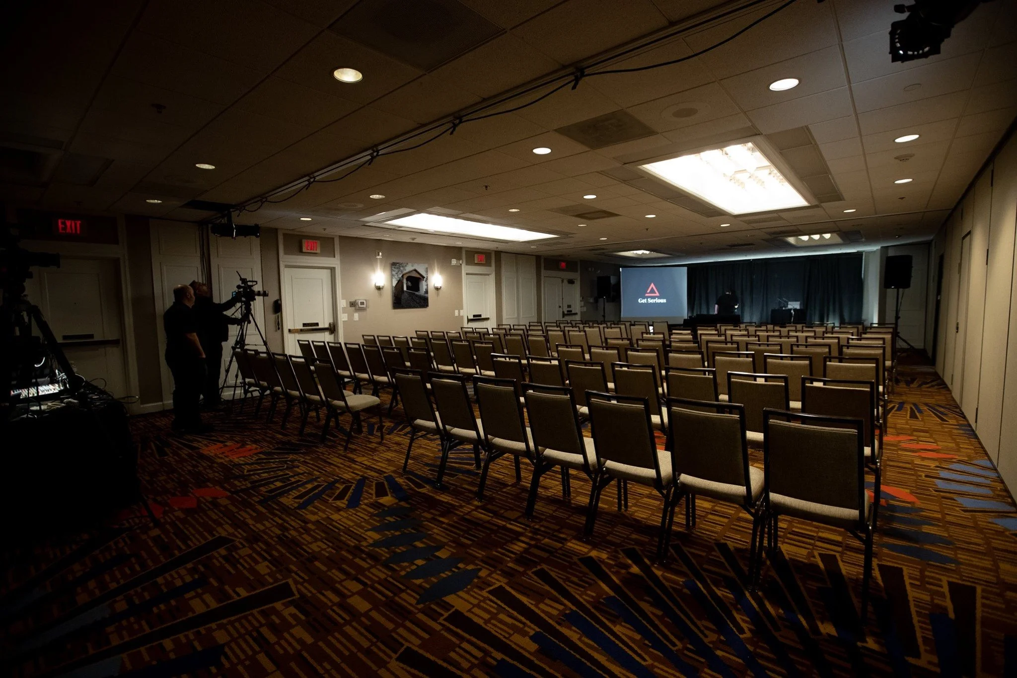 An empty conference room with rows of chairs facing a stage with a large screen displaying a red triangle and the words 'Get Serious'.