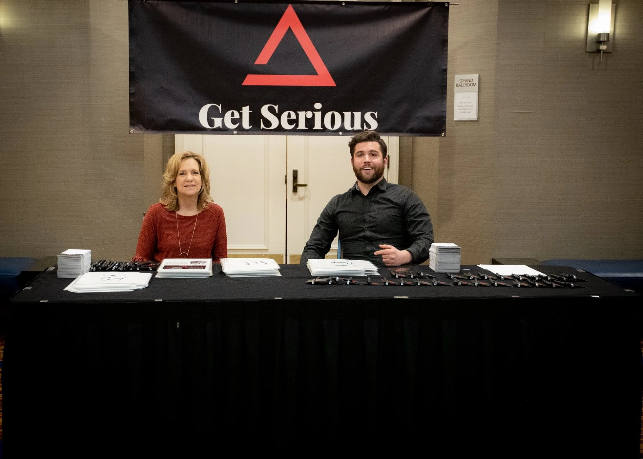 Two people seated at a registration table in front of a large sign that reads 'Get Serious' with a triangular symbol. The table has stacks of brochures and pens.
