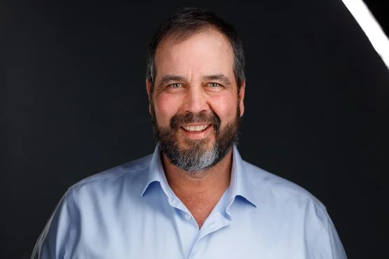 A smiling man with a beard and mustache wearing a light blue collared shirt, photographed against a black background.