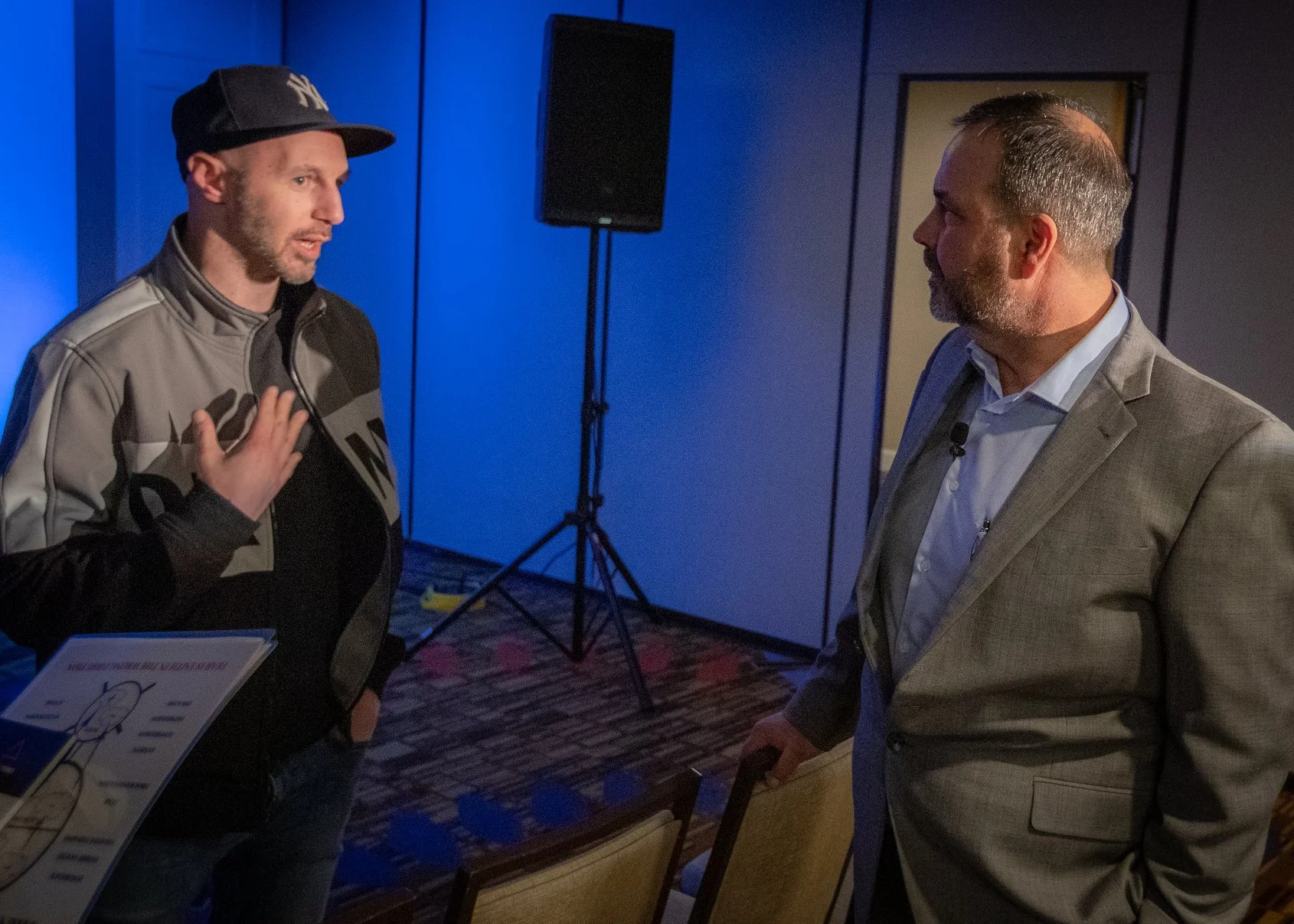 Two men face each other indoors, one in casual attire with a New York Yankees cap and jacket, speaking with a hand on his chest; the other in a beige suit with a beard, listening attentively. There are blue wall panels, a black speaker on a stand, an