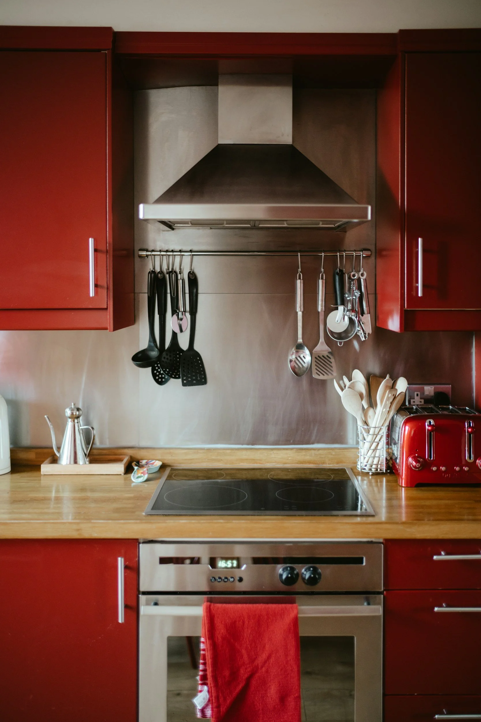 Kitchen with red cabinets, electric stove with red towel hanging, black utensils hanging on a rail, silver vent hood, red toaster, and wooden countertop.