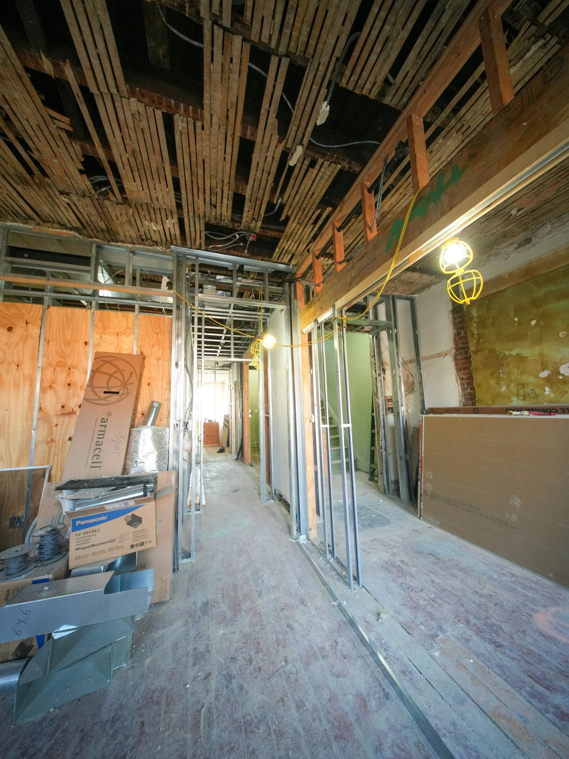 Interior of a building under construction with metal framing, exposed wooden ceiling, construction tools, and yellow temporary lighting.