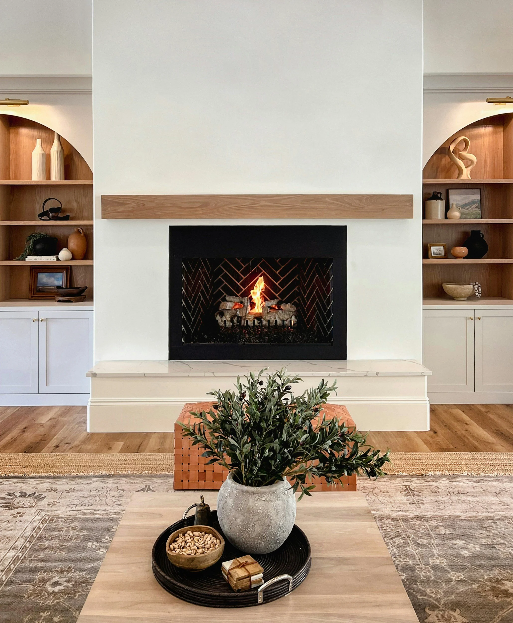 Living room with a fireplace, built-in bookshelves on either side, decorated with vases and framed pictures, and a plant in a large pot in front of a woven ottoman.